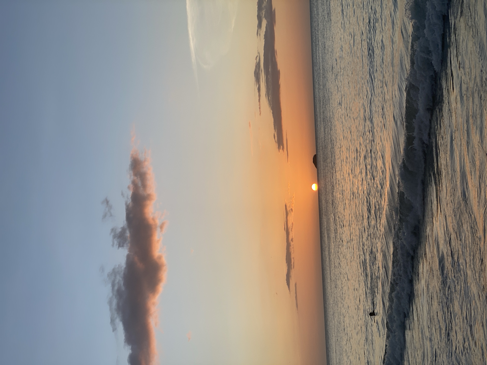 This image captures a sunset over the ocean, viewed from the beach in Pedro González, Venezuela. The sky displays a gradient of colors, transitioning from pale blue at the top to warm orange hues near the horizon, with wisps of pinkish-brown clouds. A small, dark island is visible on the horizon to the left of the setting sun. The water is calm, with gentle waves rolling onto the shore. In the mid-ground, a person is swimming, with only their head and shoulders visible above the water. The foreground shows the breaking waves and the wet sand of the beach.