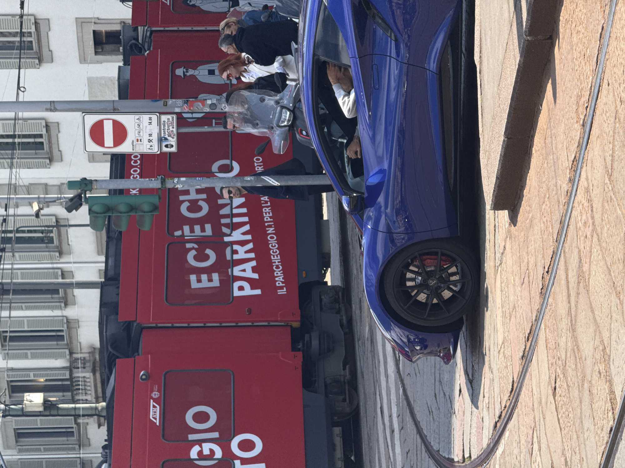 The image shows a blue car parked on the side of a street in Milan, Italy. Next to the car is a red tourist bus with the text "GIO TO" and "E C'È PARKING O" written on it. A no entry sign, along with a sign indicating time restrictions for parking, is seen near a traffic light. People can be observed inside the bus. The road is made of cobblestone and a tram track is adjacent to the sidewalk. A building with multiple windows and shutters is visible in the background.