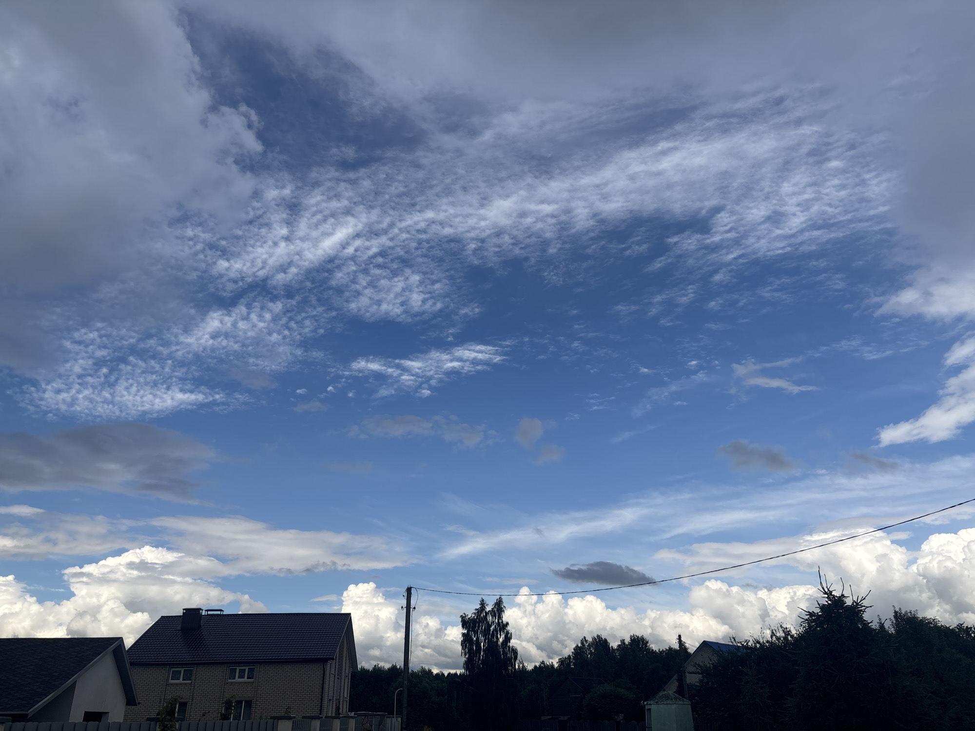 The image depicts a wide-angle view of the sky above a rural landscape in the vicinity of Капыльскі сельскі Савет, Belarus. The sky is a prominent feature, dominated by a vibrant blue expanse interspersed with various cloud formations. High in the sky, wispy cirrus clouds create delicate patterns, while lower down, more substantial cumulus clouds, some appearing bright and fluffy, and others darker and more ominous, are visible. A thin, dark wire runs horizontally across the mid-section of the frame, likely a utility line.
Below the sky, the landscape is characterized by a dense line of dark green trees forming a forest. In the foreground, several houses are partially visible. To the left, a portion of a house with a dark roof and a lighter colored facade is seen. Next to it, a larger house with a dark, pitched roof and brick walls is prominent. This house has several windows and a chimney. To the right, more trees obscure the view, but a glimpse of another building with a dark roof and a light-colored structure, possibly a shed or outbuilding, can be seen peeking through the foliage. A single tall, slender pole with what appears to be electrical insulators stands near the center-left of the frame, adjacent to the brick house. The overall scene suggests a partly cloudy day in a semi-rural area. There are no people or animals visible in the image.