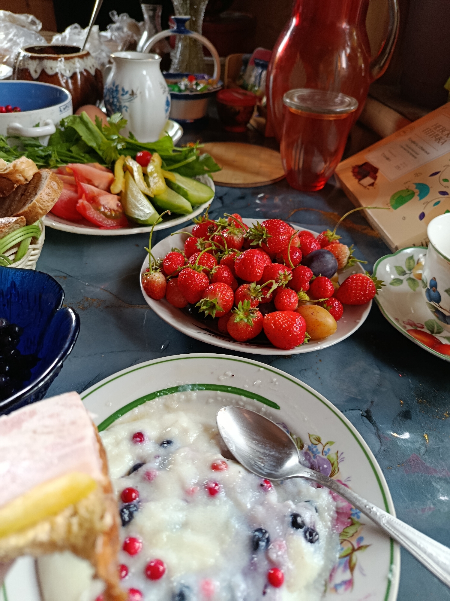 An overhead medium shot captures a rustic breakfast spread on a dark, textured surface, possibly a table, in an indoor setting. In the foreground, a slice of cake with a pale yellow layer sits on the left. To its right, a white plate with a green rim features a portion of oatmeal or porridge studded with blueberries and red currants, with a silver spoon resting on it. Behind this, a white plate with a floral design holds a generous pile of fresh strawberries, a plum, and a small yellow fruit, likely a mini-potato or apricot. A deep blue bowl filled with dark berries, possibly blueberries or blackcurrants, is positioned to the left of the strawberry plate. Further back, a plate of sliced tomatoes, cucumbers, and pickled peppers is visible, accompanied by bread slices in a woven basket. Various pitchers, a jug, and decorative vases are scattered across the background, some with reflective surfaces. The overall scene evokes a sense of a leisurely, homemade meal.