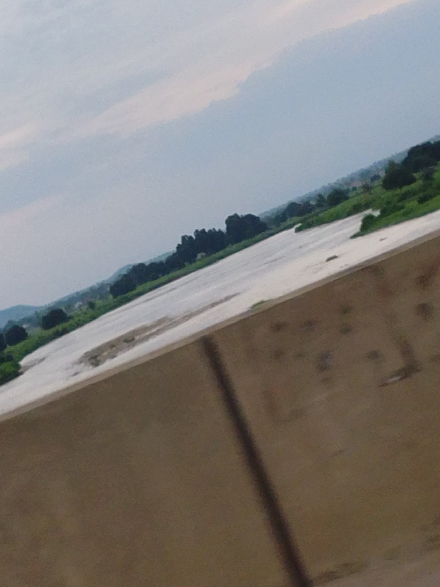 The image shows a landscape view of a river with trees and hills in the background. The foreground is dominated by a wooden surface, possibly a table or a deck, which is tilted. The sky is overcast with clouds. The scene appears to be located in a rural or semi-rural area. Specifically, this scene depicts a portion of the River Benue in Jalingo, Nigeria. The river is wide and appears to be shallow in some areas, with visible sandbanks. The surrounding vegetation is lush and green, indicating a tropical environment. The presence of hills in the distance suggests a varied topography. The wooden structure in the foreground creates a sense of depth and perspective, leading the viewer's eye towards the river and the landscape beyond.