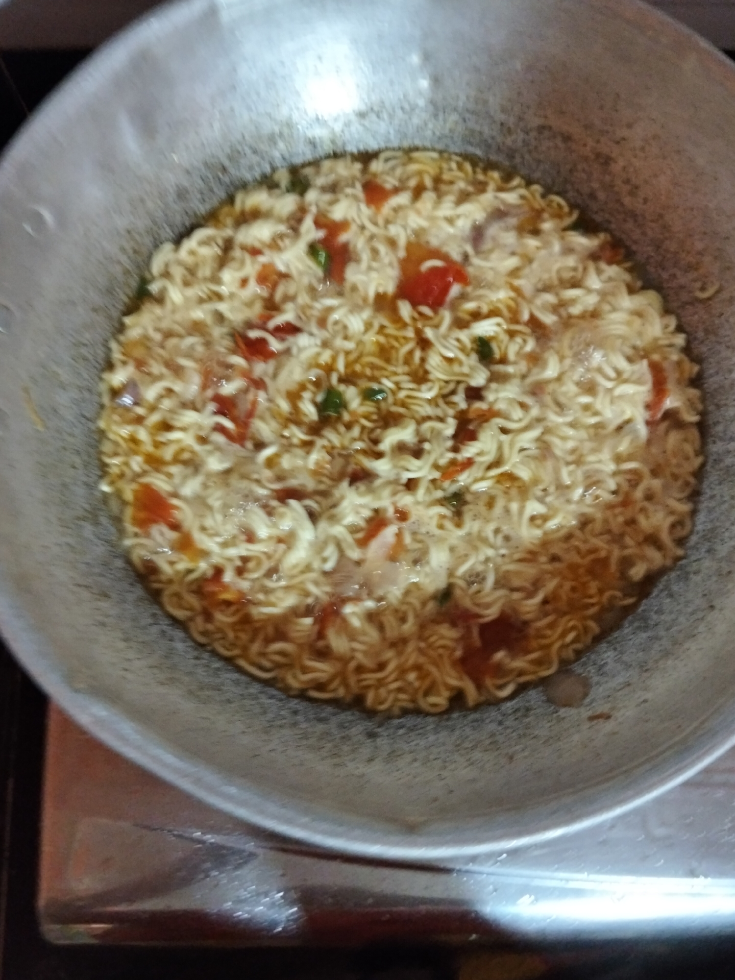 A top-down view shows a close-up of instant noodles being cooked in a wide, shallow pan. The noodles are coiled and submerged in a golden-brown broth, interspersed with chunks of red tomato and small green pieces, likely herbs or spices. The broth appears to be boiling, with small bubbles visible on the surface, indicating the cooking process is active.
The pan is metallic, with a slightly worn texture, suggesting it's well-used. It rests on a reflective surface, possibly a stovetop, which shows some smudging and a slight sheen from the lighting. The overall scene is domestic and suggests a meal being prepared. There are no people visible in the frame, but the activity implies human presence.
The lighting is overhead and somewhat harsh, casting reflections on the pan and the stovetop. The colors are dominated by the warm tones of the noodles and broth, contrasted with the cool metallic gray of the pan. The image appears to have been taken indoors, in a kitchen environment, likely during the daytime given the brightness. There is no visible text in the image. The location context of Payagaboh, Indonesia, adds a cultural layer to the domestic scene of preparing instant noodles.