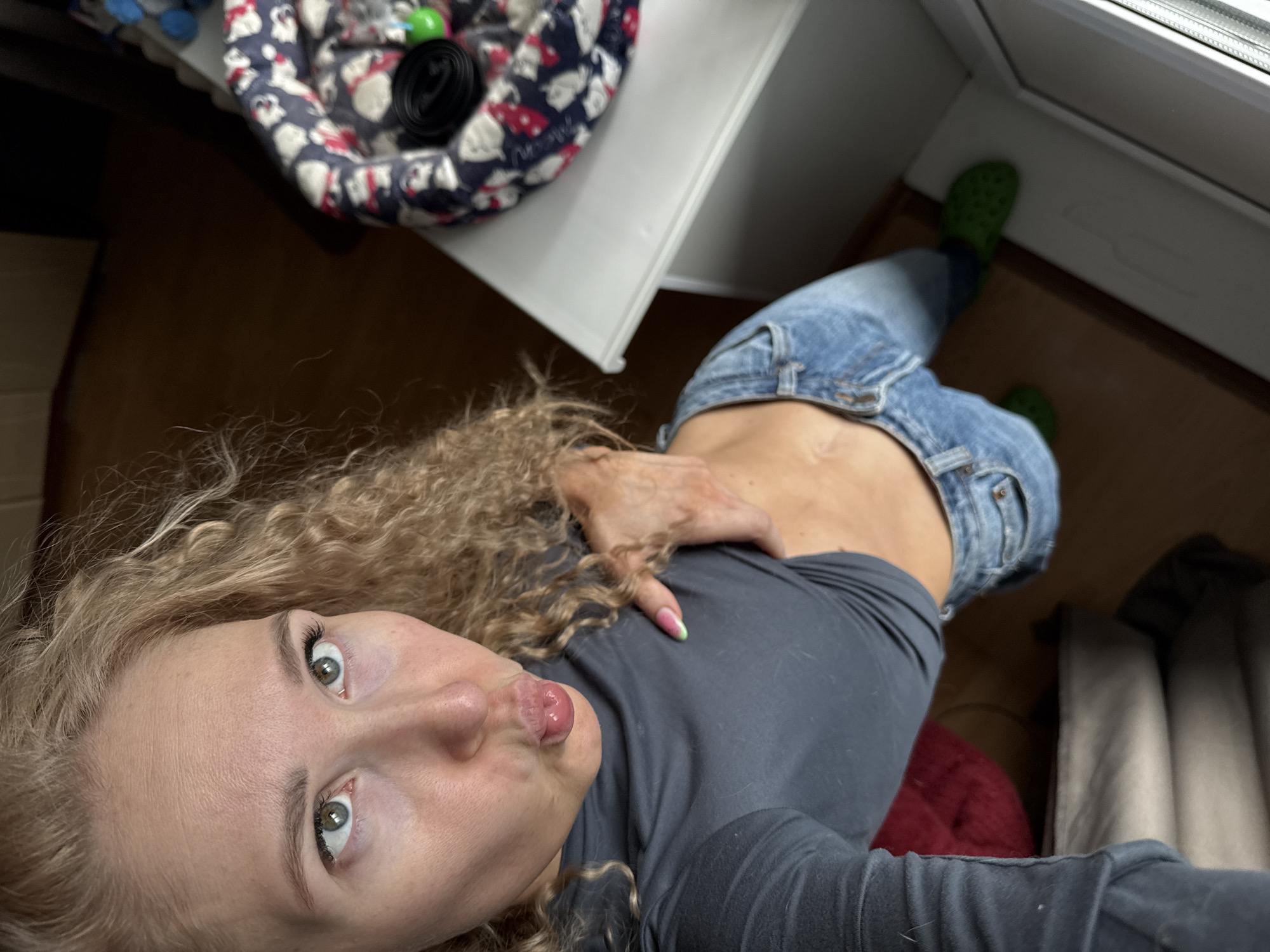 A woman with blonde curly hair, wearing a gray long-sleeved shirt and jeans, is taking a selfie. She has light skin, green eyes, and her lips are puckered. She is wearing green Crocs.
A pet bed with a pattern of mice is visible in the upper left of the frame, near a white desk or shelf. The background includes a window, brown flooring, and items of clothing.
The location is likely indoors, but there is no specific information or context to confirm if this image was taken in Perm, Russia.