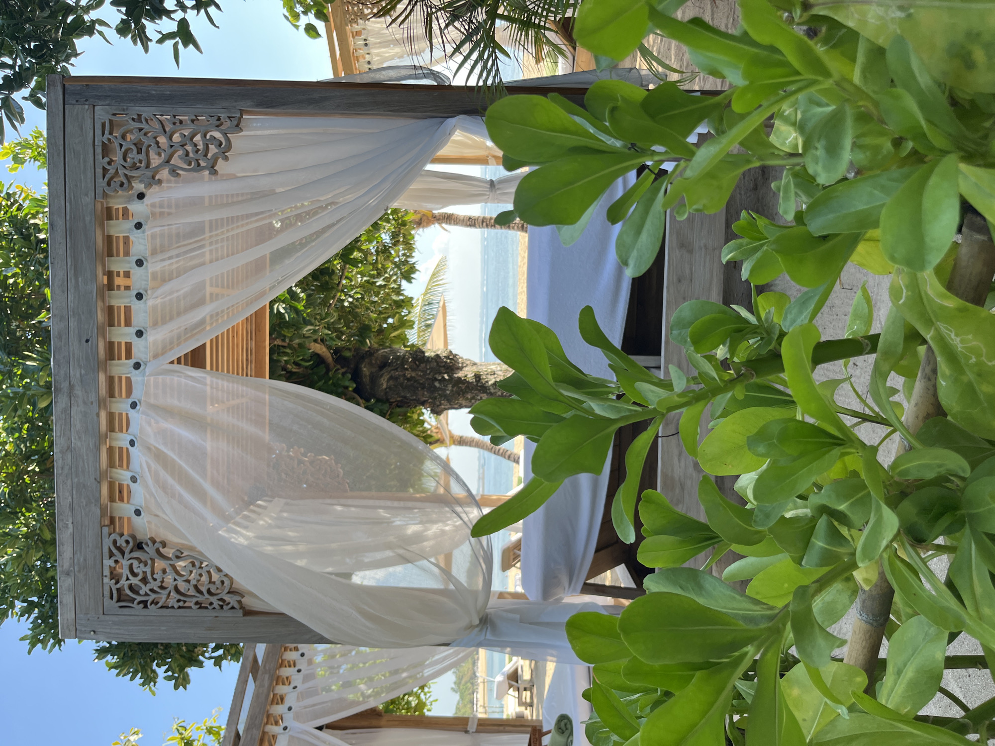 In Kuta Selatan, Indonesia, this image captures an outdoor massage cabana situated on a sandy beach. The cabana is constructed from weathered wood and features white sheer curtains that partially obscure the view of the interior. Inside the cabana, a massage bed, covered with white linens, is visible. In the background, the ocean stretches to the horizon under a clear blue sky, with a few palm trees lining the shore. In the foreground, lush green foliage partially obstructs the view of the cabana and the beach. The scene conveys a sense of relaxation and tranquility, characteristic of a tropical spa setting.