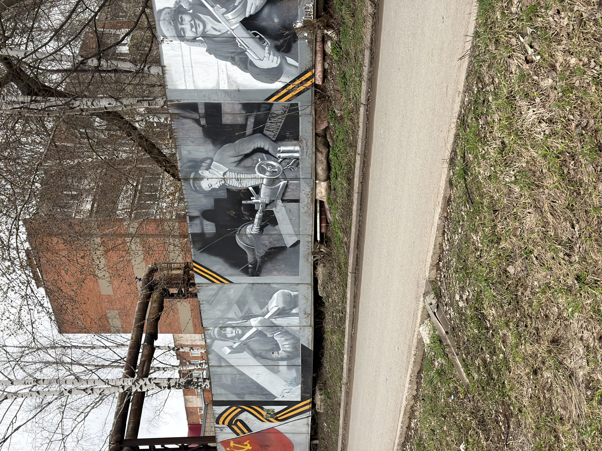 Here is a description of the image:
The image shows a series of concrete panels painted with black and white portraits of figures, appearing to be from Soviet-era propaganda. The panels are decorated with black and orange striped ribbons, a common symbol of Russian military valor. At the bottom is a painting of the Soviet flag with a hammer and sickle.
The panels are situated next to a concrete path, with an area of grass and dry leaves beyond. In the background, there's a brick building partially obscured by birch trees with bare branches.
Based on visual information and common knowledge, this is likely a commemorative mural or display. While I cannot confirm the specific location in Votkinsk, Russia without more information, it is possible the mural is located near a historical site or public space.