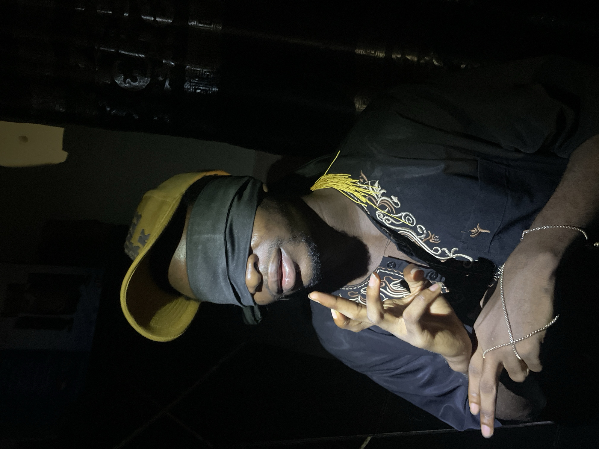 An African man with a dark complexion is lying with his back facing the camera. He is wearing a dark top with gold embroidery and a yellow hat covered in dark fabric over his eyes. He is wearing a silver chain bracelet on one hand. The location is Lokoja, Nigeria, but the specific context or environment within Lokoja is not identifiable from the image.