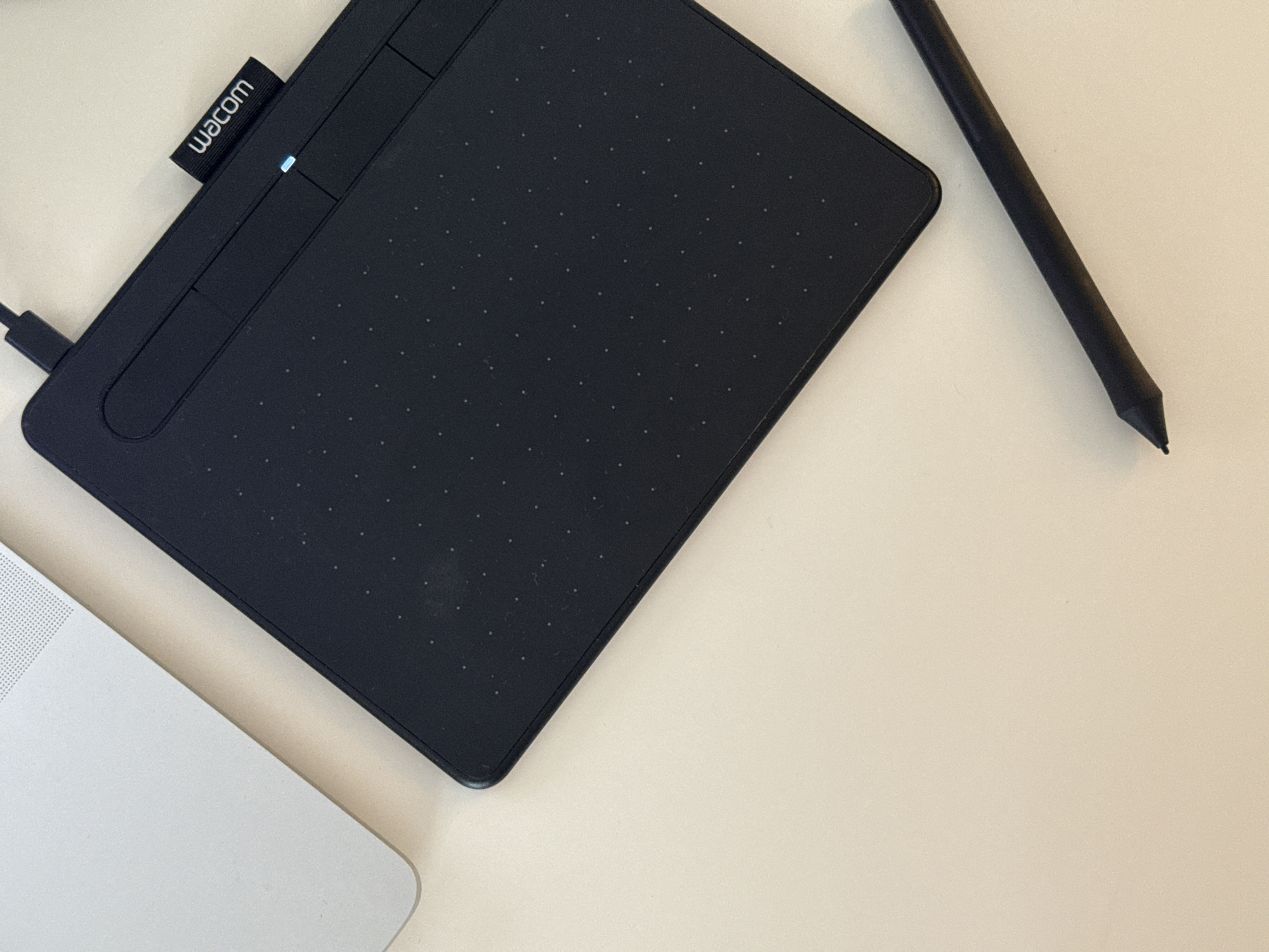 A high-angle, close-up shot depicts a Wacom graphics tablet situated on a neutral-toned surface. The tablet is black, rectangular with rounded corners, and features a textured surface of tiny dots. A Wacom logo is visible on the upper left corner, along with a small blue light indicating power. A black stylus is positioned to the right of the tablet. The lower left corner of a light-colored laptop is visible, partially obscured by the tablet. The background is a uniform, light beige, suggestive of a desk or tabletop.
Location information (Dikaya Gar', Russia) is irrelevant to the image's content.