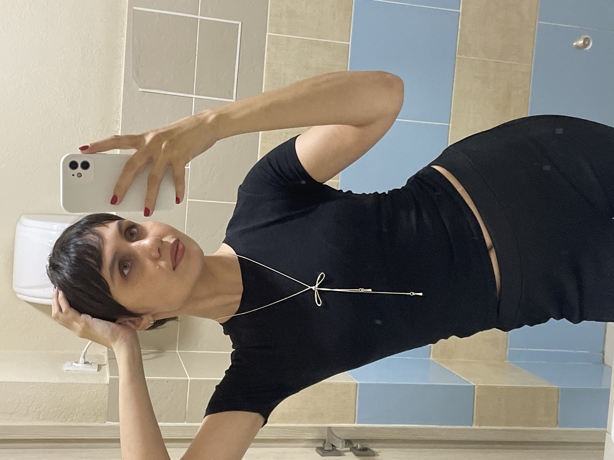 A person is taking a selfie in a bathroom. The person has short, dark hair and is wearing a black shirt and necklace. The bathroom has beige and blue tile walls. A phone is in the top left of the frame. A toilet is in the background. The location is not specified as being in the city of Izhevsk, Russia.