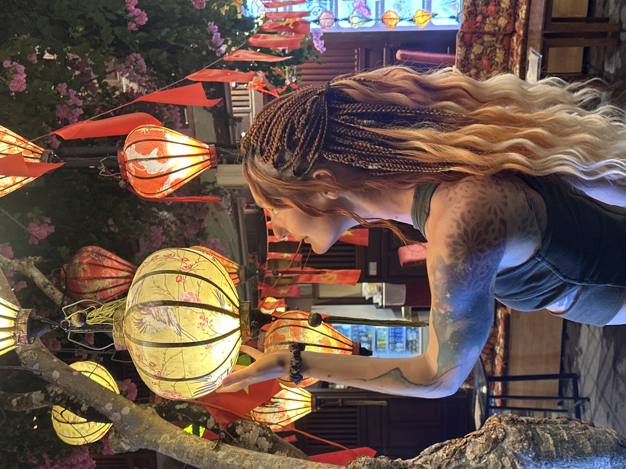 In a close-up shot, a woman with braided ginger hair and a dark tank top is seen from the chest up, turned to her right. Her arm is extended, reaching towards a cluster of illuminated lanterns. The lanterns, in shades of yellow and orange, are decorated with intricate patterns of birds and flowers. They are suspended from tree branches, alongside red triangular flags that add a festive touch. The background suggests an outdoor setting, possibly a street or market, with glimpses of buildings and foliage. The lighting implies it is evening or night. The woman's expression is one of gentle curiosity as she interacts with the lanterns. Her arm is adorned with a colorful, starry tattoo. The overall scene evokes a sense of cultural immersion and enchantment, likely captured during a festival or a visit to a vibrant night market in Hội An, Vietnam.