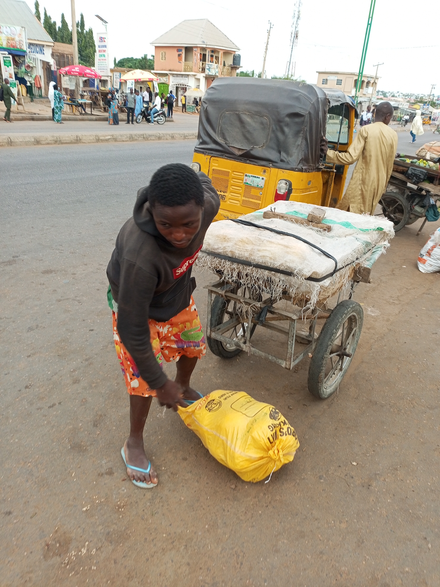 In Tempure, Nigeria, a young boy wearing a dark hoodie with a red logo and orange shorts with colorful prints is actively engaged in moving a large yellow sack on a paved street. He is seen leaning down, gripping the sack with both hands and pulling it along. He is wearing light blue flip-flops. Behind him, a cart is loaded with several large sacks covered by a textured fabric, secured with black straps. A portion of a yellow auto-rickshaw is visible to the left of the cart. Further in the background, other individuals and vehicles are present along the street, with various buildings and shops lining the area. The overall environment suggests a bustling market or roadside setting.