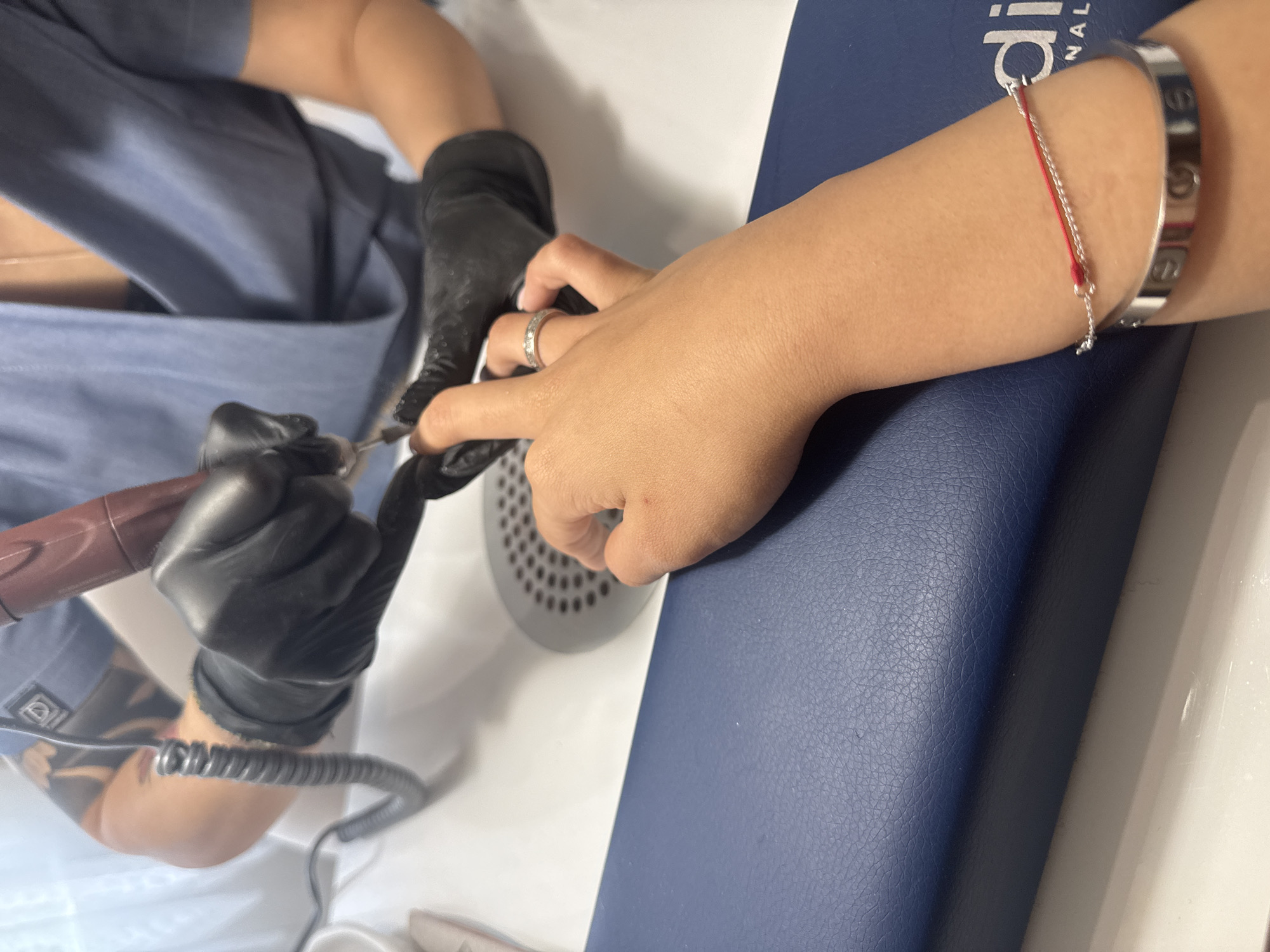 The image shows a close-up of a manicure session. A person's hand with a ring on their finger and a bracelet on their wrist is resting on a blue padded surface. Another person, wearing black gloves and a blue uniform, is using an electric nail file on the nails of the hand. The background shows a white surface with a sink and a drain. The scene appears to be in a nail salon. No specific location in Plovdiv, Bulgaria can be determined from the image.