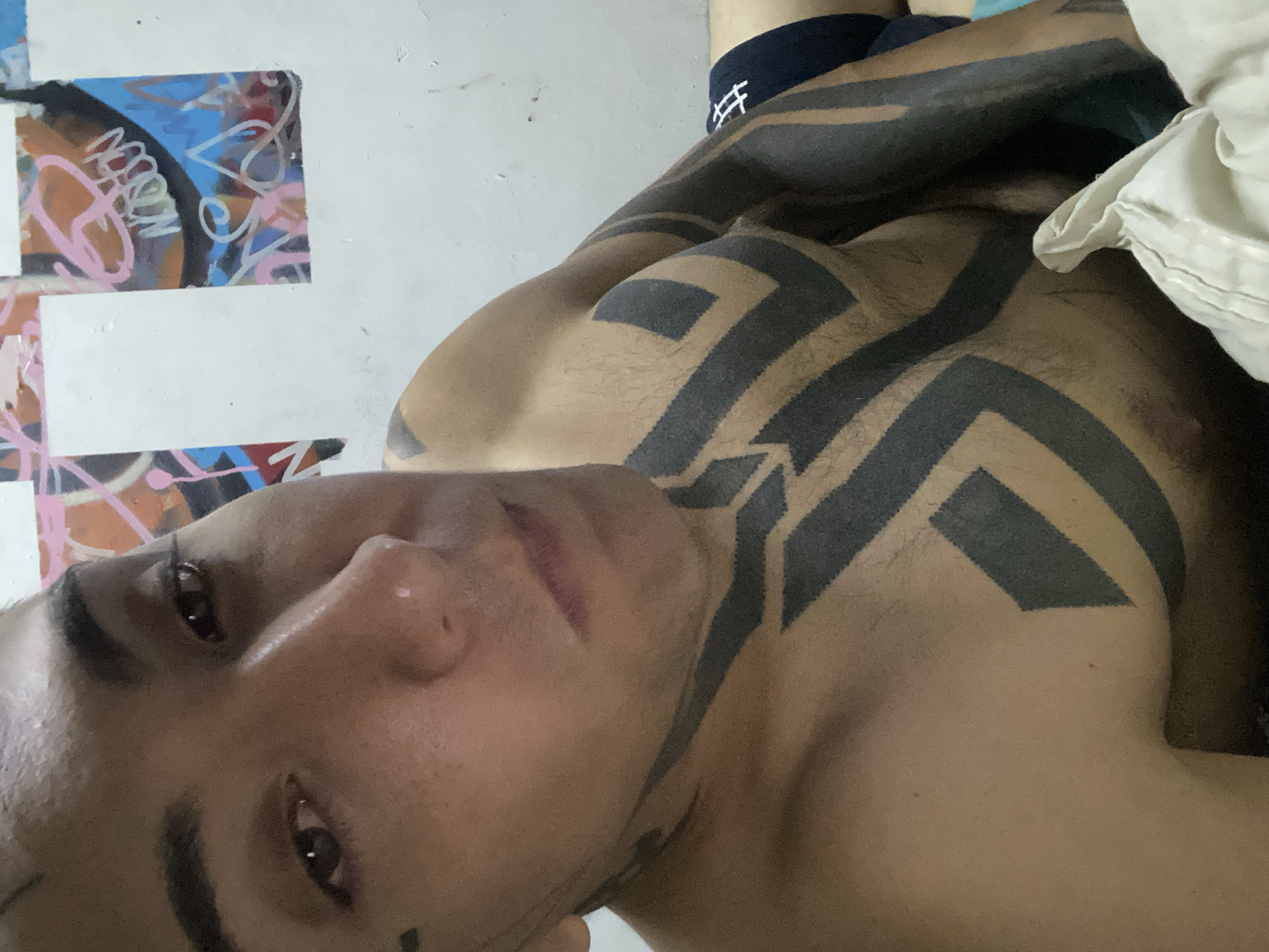 A close-up, low-angle shot features a shirtless individual with extensive black geometric tattoos covering their chest, shoulders, and arms. The person is lying down, and their face is prominently displayed, showing dark hair, brown eyes, and an intent gaze. The background includes a white wall with graffiti-style art in various colors (pink, blue, orange, white) and a blue pair of underwear. The scene appears to be indoors, possibly a bedroom. There is no evidence to indicate the location is Buenos Aires, Argentina.