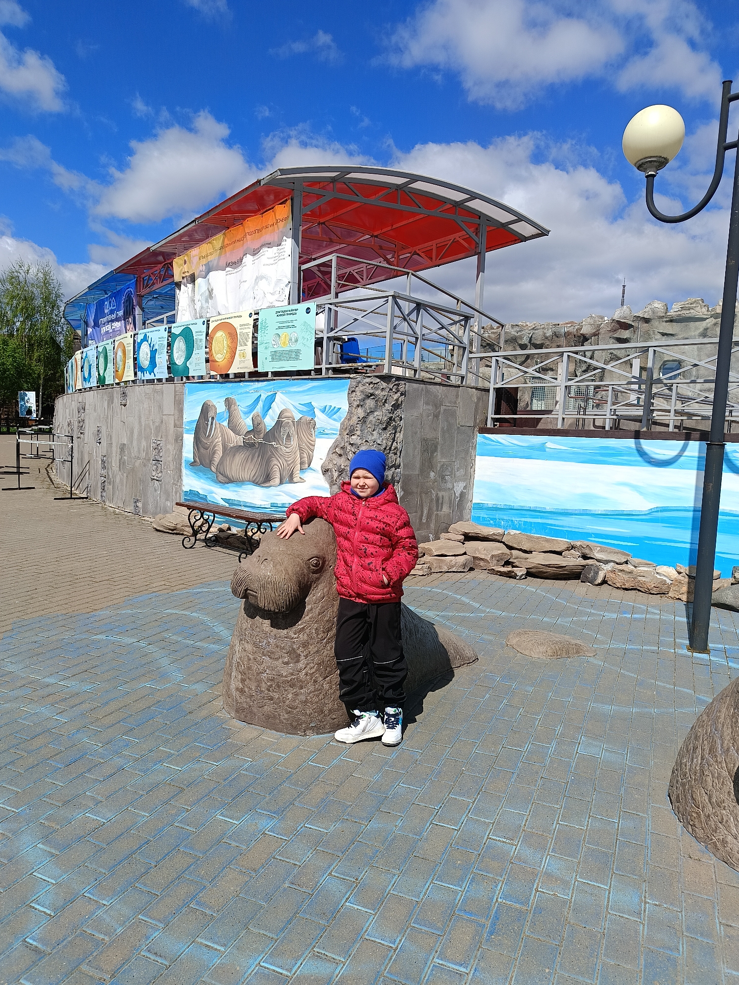 A child stands next to a walrus statue in the city of Izhevsk, Russia. The child, wearing a red jacket and blue hat, rests their hand on the head of the brown walrus statue. The statue sits on a blue brick walkway. Behind the statue is a concrete wall decorated with a walrus mural and informational signs. Above the wall is a structure with a red roof and metal framework. A light pole is visible to the right. The sky is blue with scattered clouds.