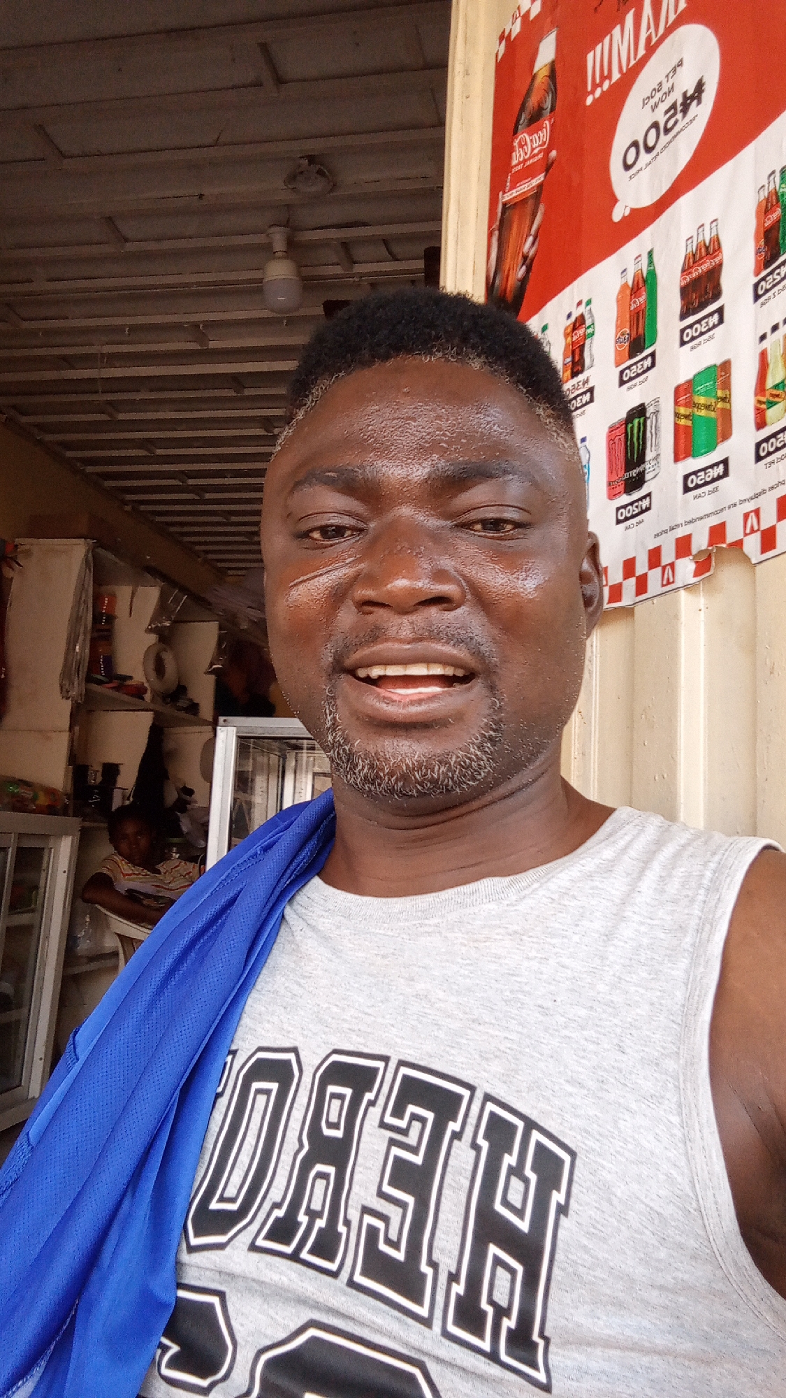 This is a close-up selfie of a dark-skinned man smiling broadly, with a salt-and-pepper beard and short, dark hair that is lighter at the temples. He appears sweaty, indicative of warm or humid weather. He is wearing a light grey sleeveless t-shirt with "HERO" printed in large, black outlined letters across the chest (appearing reversed in the image). A blue garment is draped over his left shoulder.
The setting is an indoor space, likely a small shop or market stall in Sabon Tasha, Nigeria, during daylight hours. In the background to the man's left, shelves stocked with items are visible, along with a light fixture hanging from a wooden-slat ceiling. A child is partially visible sitting in a chair further back. To the man's right, a red and white banner prominently displays advertisements for beverages, including Coca-Cola and Monster Energy.
Visible text on the banner includes reversed prices like "₦200", product names such as "MISOO" and "WOW", and phrases like "pet bottle". Other reversed text seems to be promotional details. The man's facial expression conveys happiness or contentment.