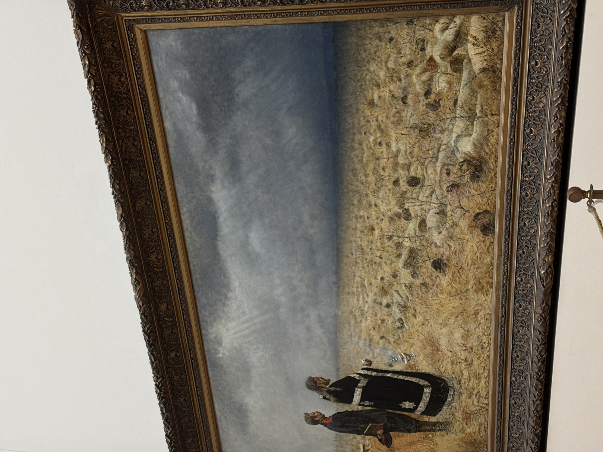 A framed painting depicts a landscape scene with a cloudy sky taking up the left half of the painting. The right half of the painting depicts a field of dry grass and sparse vegetation. In the foreground, two figures stand: one wearing a black robe with silver accents and holding an object that appears to be a smoking censer, and another figure standing slightly in front, wearing a dark coat and red shoes. The painting is encased in an ornate, dark-colored frame. This painting is called "Eternal Peace" by Isaac Levitan, which is in the State Tretyakov Gallery in Moscow, Russia.