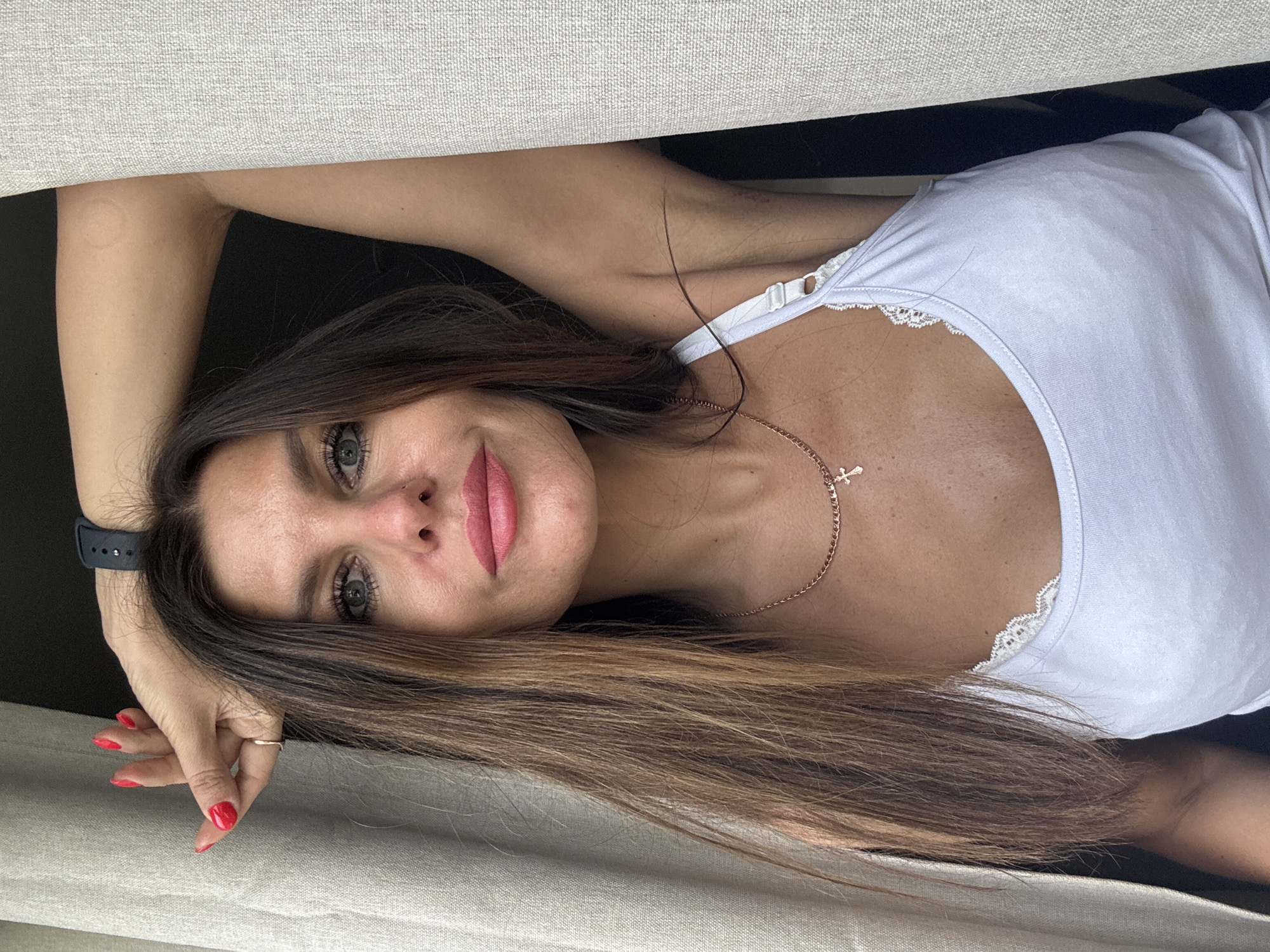 A woman with brown hair highlighted with blonde is reclining on a beige surface, possibly a couch or bed, in an indoor setting. She is wearing a white tank top with lace trim around the neckline and a delicate gold necklace with a cross pendant. Her left hand, adorned with red nail polish, is resting near her face, and she is wearing a dark-colored smartwatch on her left wrist and a gold ring on her left hand. The background is dark and indistinct. There is no information to indicate that the photograph was taken in Chaikovsky, Russia.