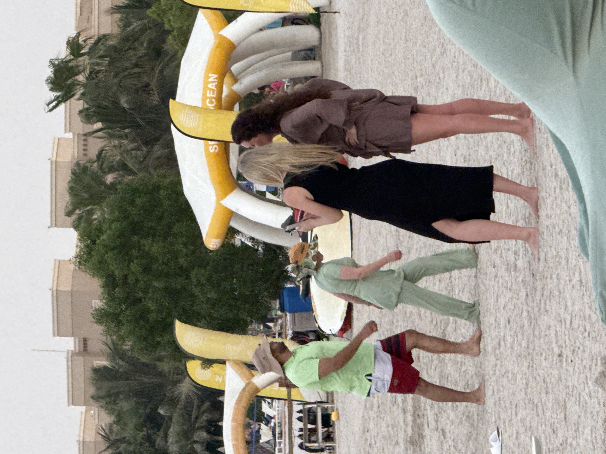 The image depicts a sandy beach with several people and structures. In the foreground, a man in a lime green polo shirt and red and white striped shorts walks on the sand, barefoot. Beside him, a woman in a light green outfit and a patterned hat also walks. In the middle ground, two women are visible. One, with blonde hair, wears a black dress and holds a phone. The other, with dark hair, wears a brown linen-like dress and has her hands on her hips. Behind them, an inflatable structure with "SEA" and "OCEAN" written on it is visible, along with yellow banners. In the background, a resort or hotel with multiple stories and light-colored facades is seen, surrounded by palm trees and other greenery. The sky is overcast. The location is identified as an unknown area in the United Arab Emirates.