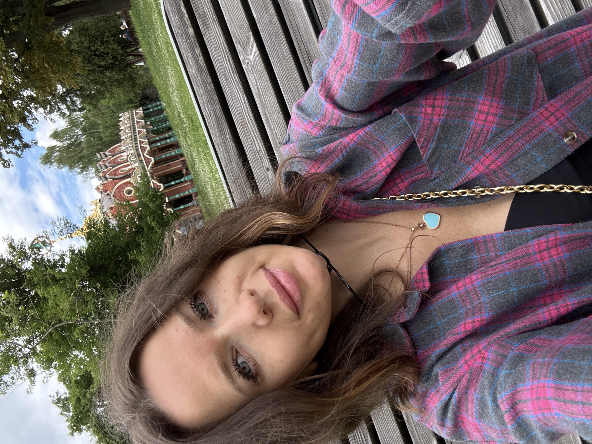A woman with shoulder-length brown hair, wearing a pink and gray plaid shirt over a black top, is sitting on a wooden bench. She is looking at the camera and smiling. In the background, there is a park with green grass, trees, and a building with red and white architectural details. The building appears to be a gazebo or folly. The sky is cloudy. The image is taken outdoors. The building in the background, with its distinctive red and white decorative facade and columns, is characteristic of the architecture found in Tsaritsyno Park in Moscow, Russia, often referred to as the "White Hall" or "Palace of Tsaritsyno".
