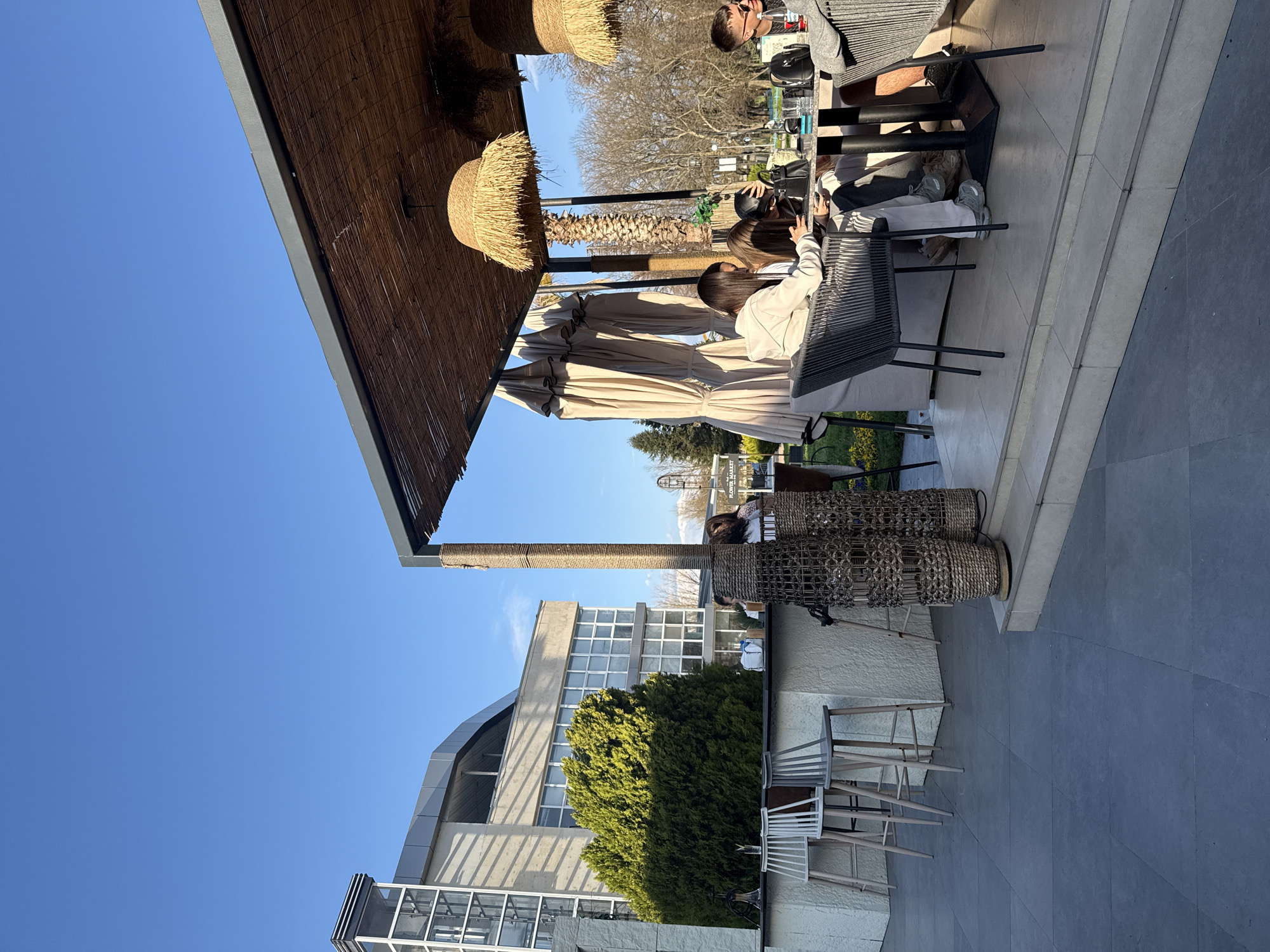 The image depicts an outdoor cafe setting in Plovdiv, Bulgaria. Patrons are seated at tables under a partially covered area with straw-like lamps hanging overhead. The cafe is next to a building with a curved roof and a large green bush. The sky is clear and blue. In the background, trees and possibly mountains can be seen. People are interacting at the tables, engaged in conversation and using electronic devices. Wicker chairs and tables are visible. The ground is paved.
