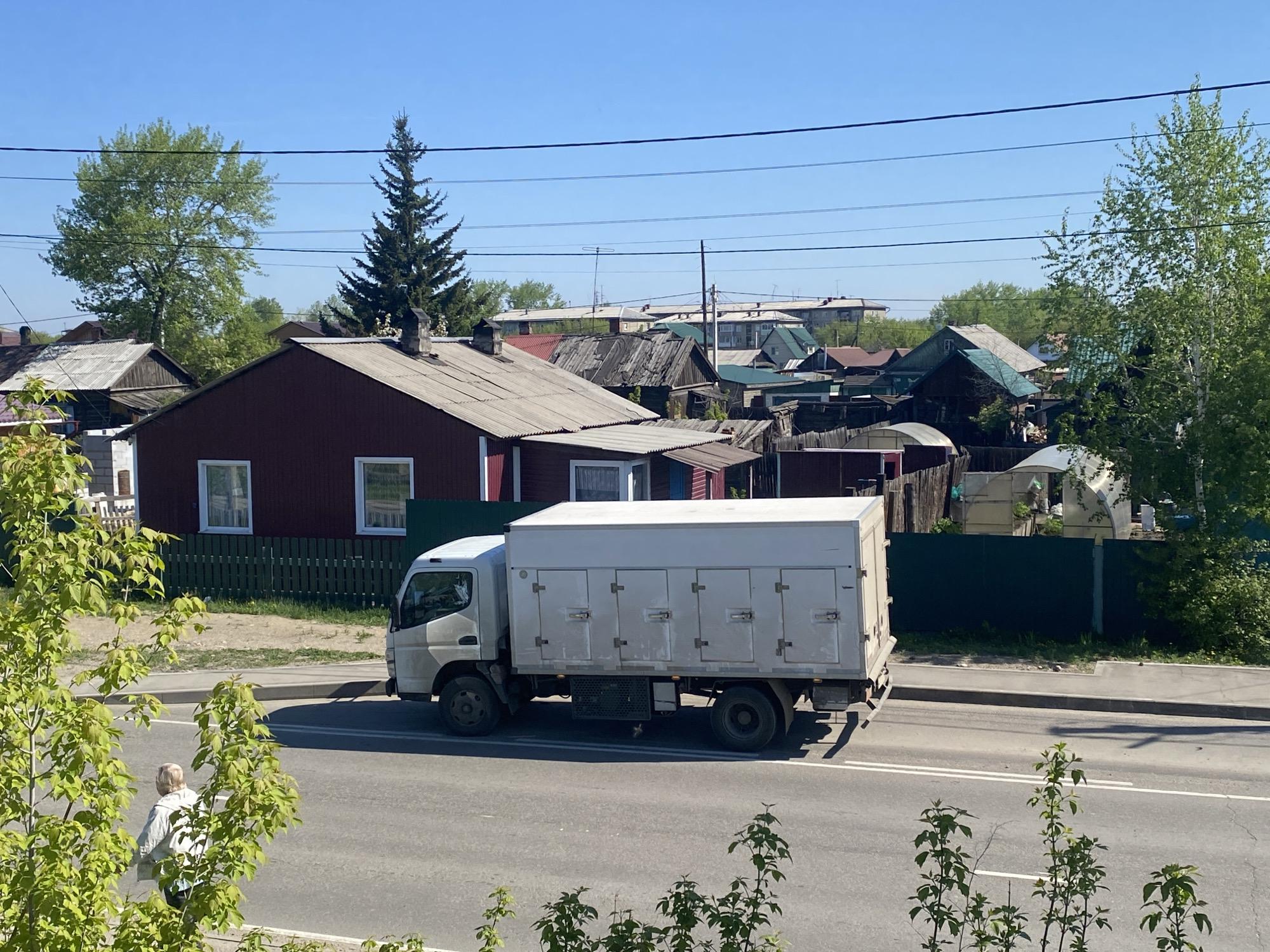 The image depicts a street scene in Angarsk, Russia on a sunny day. A white delivery truck with a multi-door box body is traveling on the asphalt road. To the left of the truck, an elderly person with short, light hair is walking on the sidewalk. Background houses with varying roof structures and colors are visible behind the fence, with assorted trees and greenery. Electrical wires run across the sky.