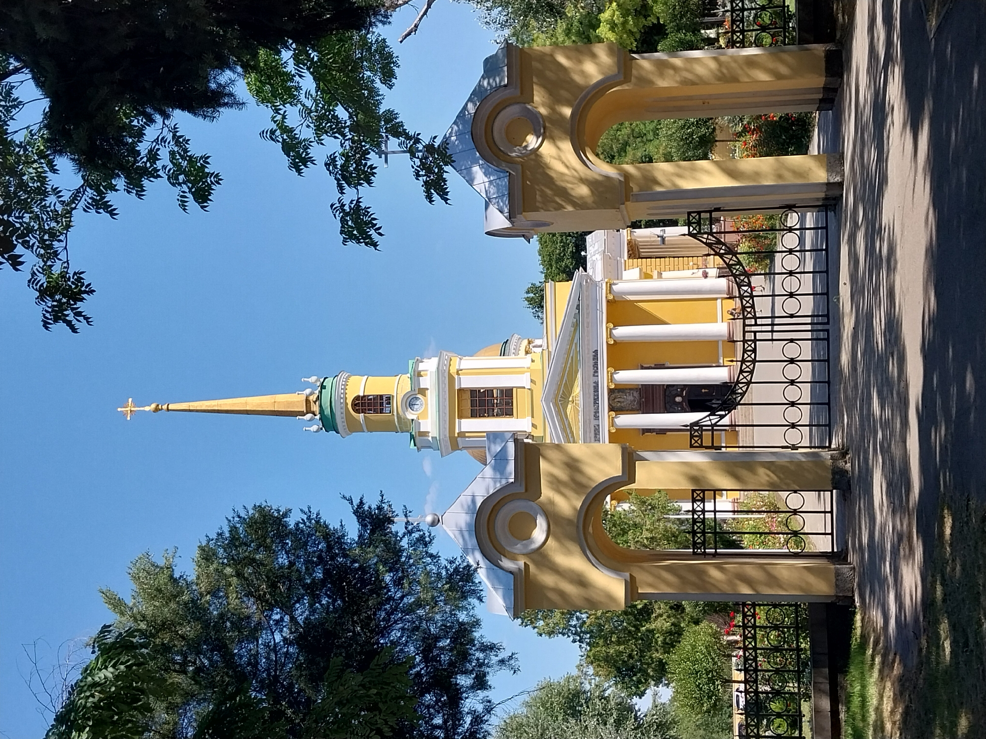 The image shows the Transfiguration Cathedral in Dnipro, Ukraine, on a clear, sunny day. The cathedral is a bright yellow building with white columns and a tall, slender yellow spire topped with a golden cross. The entrance to the cathedral grounds is marked by two yellow archways with ornate black metal gates. Green trees and bushes surround the building, with some red flowers visible near the entrance. The sky is a vibrant blue. No people are clearly visible in the foreground, but a few figures can be seen through the gates in the background. The overall impression is one of a well-maintained religious site in a pleasant, sunny environment.
