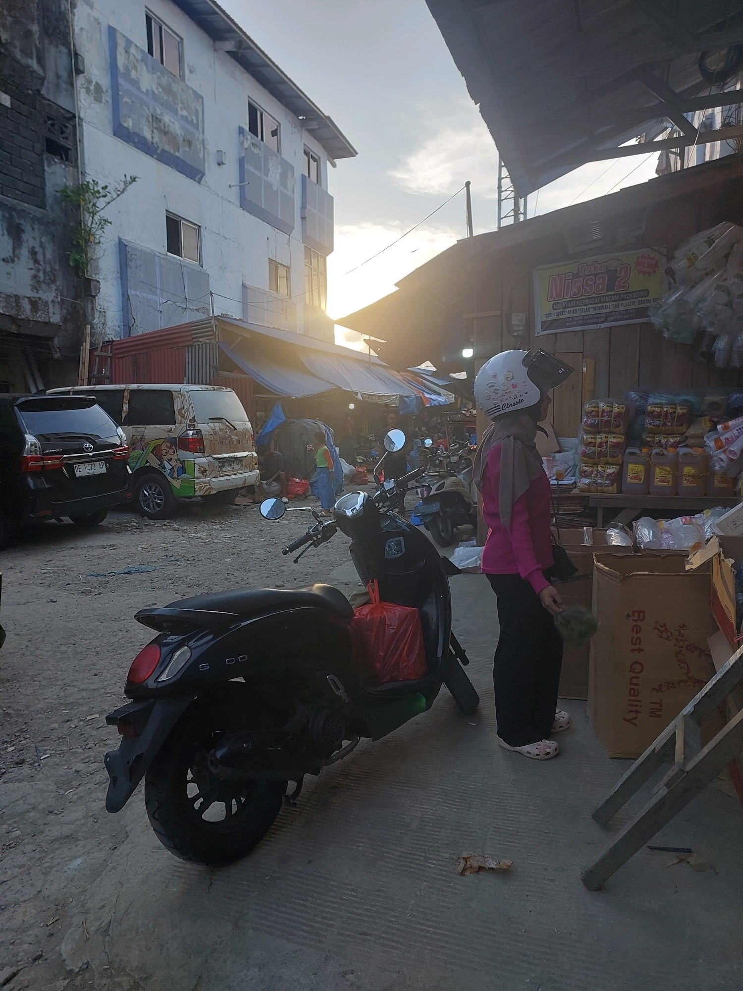 The image depicts a street scene in Ambon, Indonesia, likely a market area, given the presence of stalls and goods. In the foreground, a black scooter is parked, with a red plastic bag on its rear. To the right, a woman wearing a pink shirt, black pants, and a white helmet with stickers stands facing a stall. She is holding a green object in her right hand. Behind her, a stall displays various goods, including bottles of liquid and packaged items, on shelves and in cardboard boxes. One box prominently features the text "Best Quality". In the mid-ground, two cars are parked: a black SUV on the left and a white van decorated with cartoon characters on the right. The van has an Indonesian license plate that reads "DE 1472 AP". Further in the background, a multi-story building with white and blue accents stands to the left, with some of its walls showing wear and tear. The overall environment suggests a busy, outdoor commercial space with a mix of residential and retail elements. The time of day appears to be late afternoon or early evening, with the sun casting long shadows and a soft light across the scene.