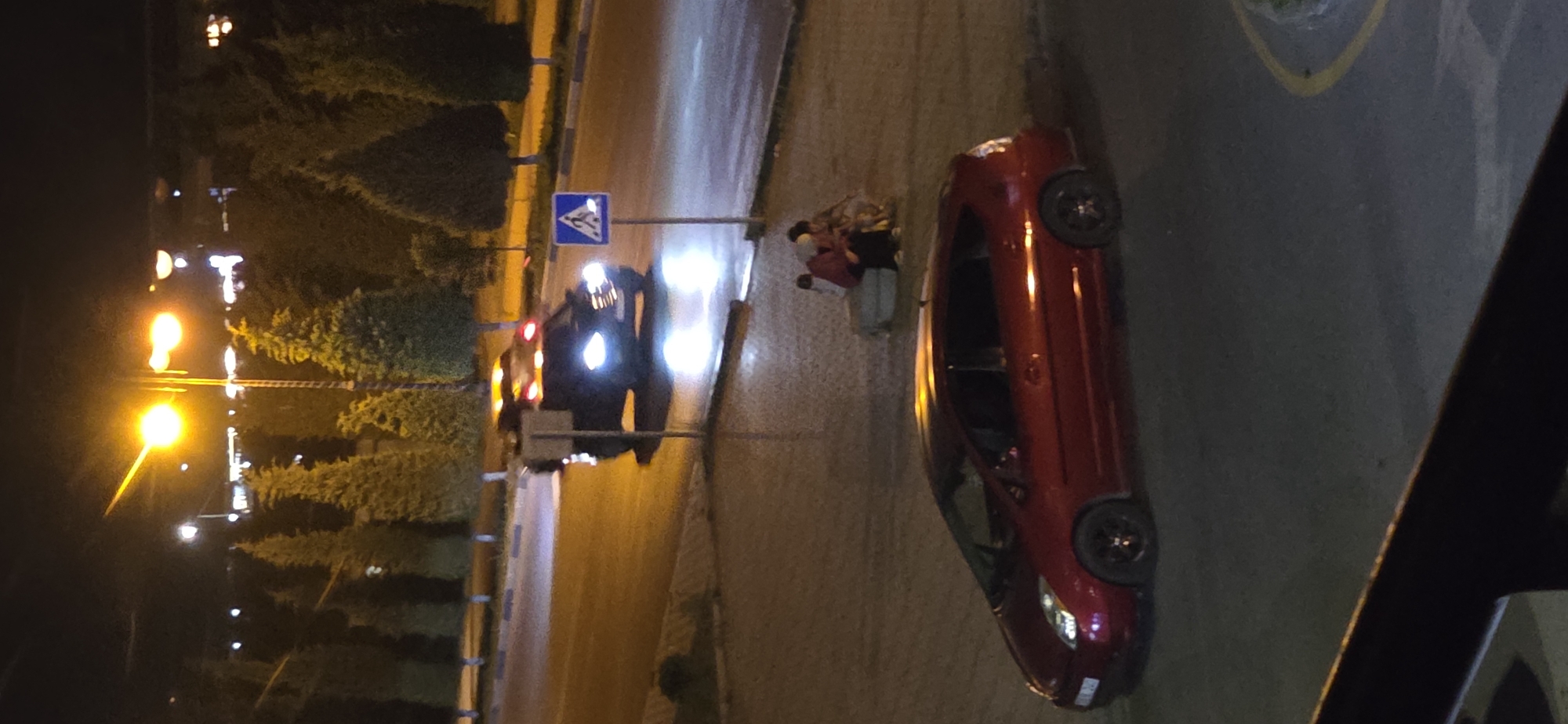 The image depicts an urban street scene at night, likely in Sumgayit, Azerbaijan, illuminated by streetlights and vehicle headlights. In the foreground, a red hatchback car is parked, facing left, with its headlights off. Further back, a dark-colored vehicle, possibly an SUV or a police car with flashing lights on its roof, is in motion on the road, its headlights on. A pedestrian crossing sign is visible to the right of this moving vehicle. A small group of people, appearing to be sitting or crouching, are gathered near a concrete structure on the sidewalk. The background shows a line of trees and distant building lights. The overall environment suggests a public area, possibly near a park or a commercial zone, with asphalt roads and paved pedestrian walkways.