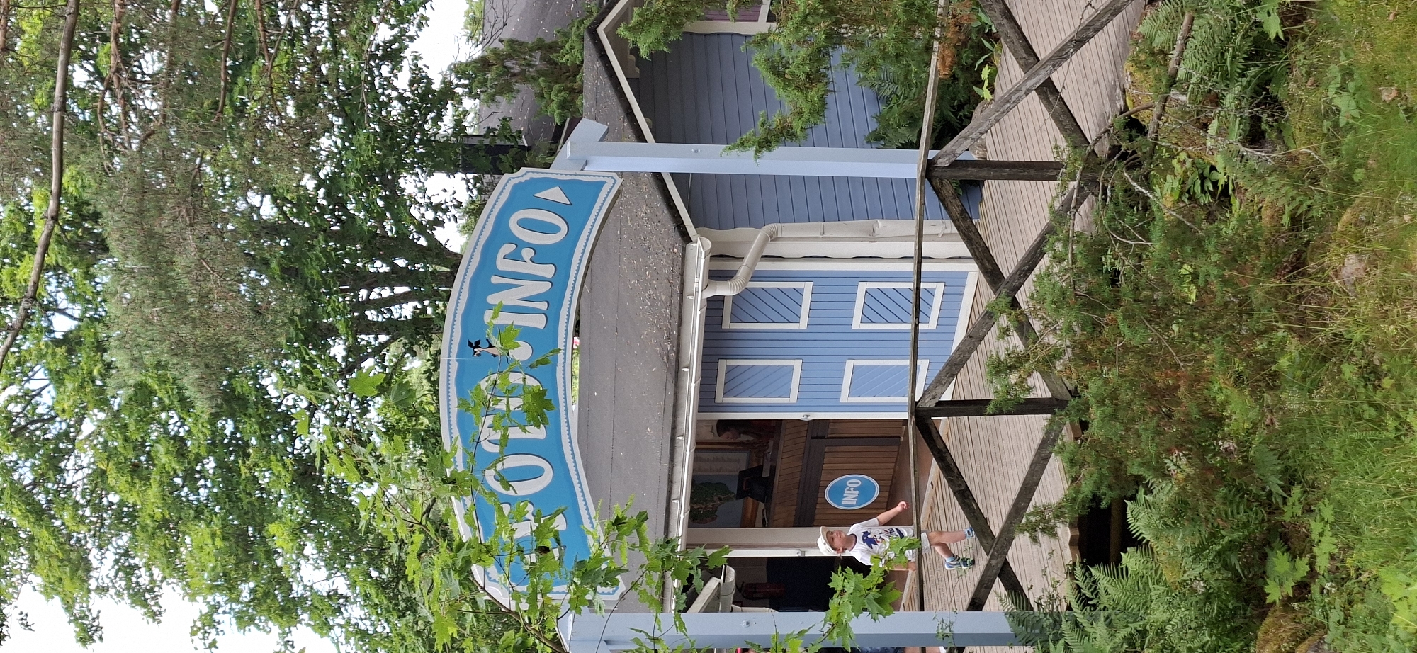 The image depicts the entrance to an information kiosk labeled "INFO" with a blue and white sign featuring a cartoonish character. The kiosk itself is painted blue with white trim around its windows and doors. A wooden walkway leads up to the kiosk, and a young boy wearing a white t-shirt, shorts, and a cap is walking along this walkway away from the kiosk. He is looking to his left. The surrounding environment is lush with green trees and foliage, suggesting an outdoor location. The presence of the "INFO" sign and the setting indicate this is likely an information center within a park or tourist attraction. Given the context of Naantali, Finland, this could be an information point within Moomin World or another recreational area in the city.
