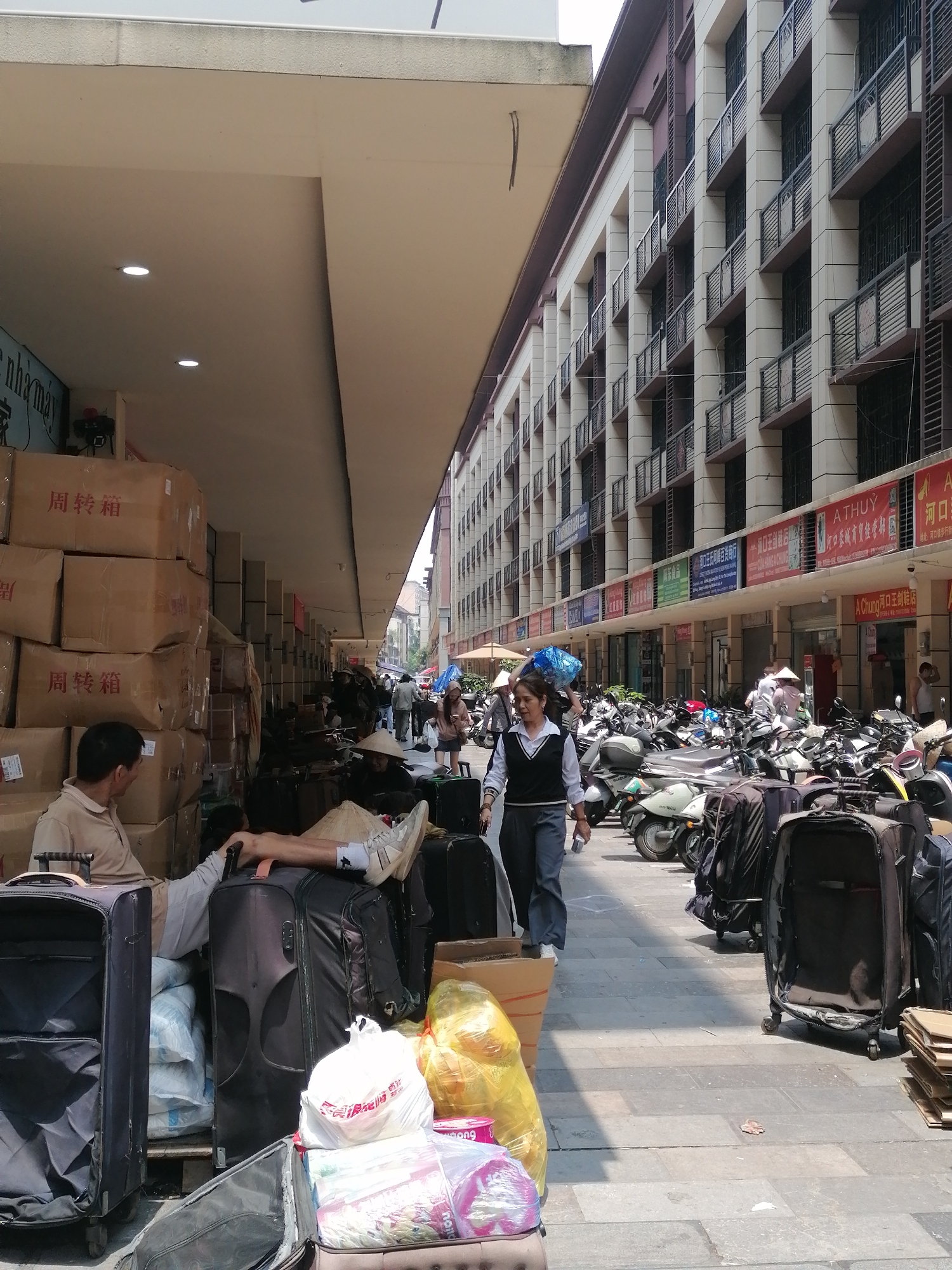 This photo was taken in the city of Hekou, China. It depicts a bustling street scene with people and parked motorbikes. The foreground shows stacks of cardboard boxes with Chinese characters, and many pieces of luggage. A man in a beige shirt sits near the boxes. One man in the middle ground has his feet up on a piece of luggage under a conical hat. A woman in a vest and jeans walks towards the camera. Behind her, people with conical hats can be seen. The right side of the image is cluttered with parked motorbikes and more luggage. Above the storefronts, signs with Chinese characters and English words ("A THUY", "A Chung") indicate businesses. The background features a multi-story building with balconies. The street is paved with rectangular tiles.