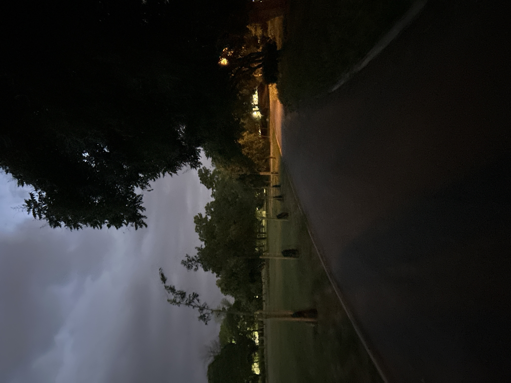 The image depicts a dimly lit park at night. A paved pathway stretches along the right side of the frame. The left side reveals a cloudy sky and silhouettes of trees. Lights illuminate the park in the distance. The location could potentially be a park in Plovdiv, Bulgaria. No people, subjects, actions, or interactions are visible.