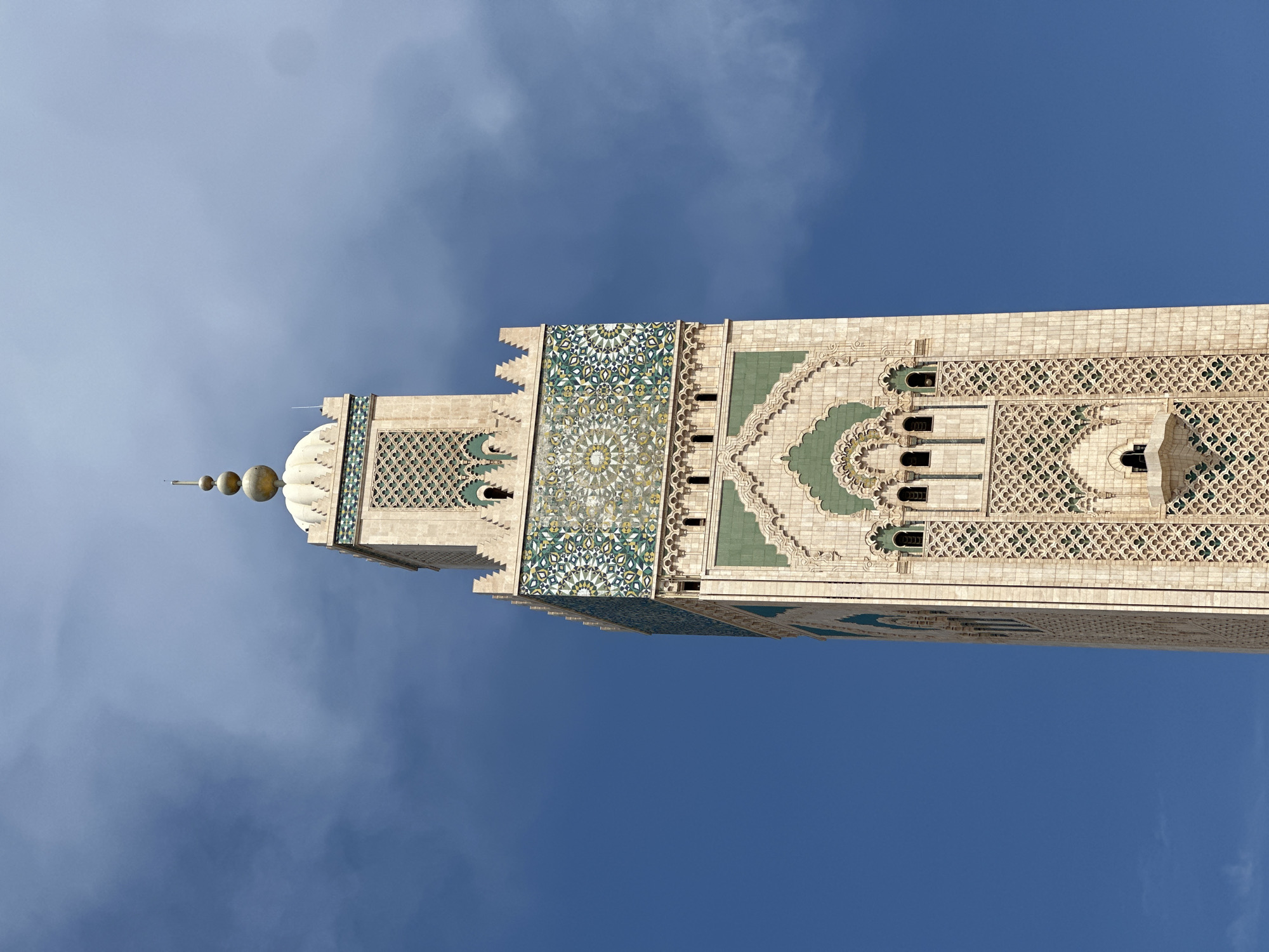 A close-up, vertical shot captures the intricately detailed minaret of a mosque against a vibrant blue sky with scattered white clouds. The minaret, indicative of Moroccan Islamic architecture, is predominantly light-colored stone adorned with elaborate geometric patterns and Zellige tilework. These patterns feature a complex interplay of green, white, yellow, and darker blue hues, showcasing radial symmetry and traditional Islamic motifs.
The structure features multiple tiers of decorative elements, including arched windows, carved stonework, and tessellated surfaces. Towards the apex, a dome-like section transitions into a series of three golden spherical finials, topped by a slender spire, characteristic of minarets in the region. The perspective is upward-looking, emphasizing the grandeur and height of the minaret.
The clear sky suggests bright daylight conditions, and the absence of shadows indicates a well-lit scene, likely during the late morning or early afternoon. The location context provided confirms this is the Hassan II Mosque minaret in Casablanca, Morocco, renowned for its monumental scale and rich ornamentation. No human figures or discernible text are visible in the image.