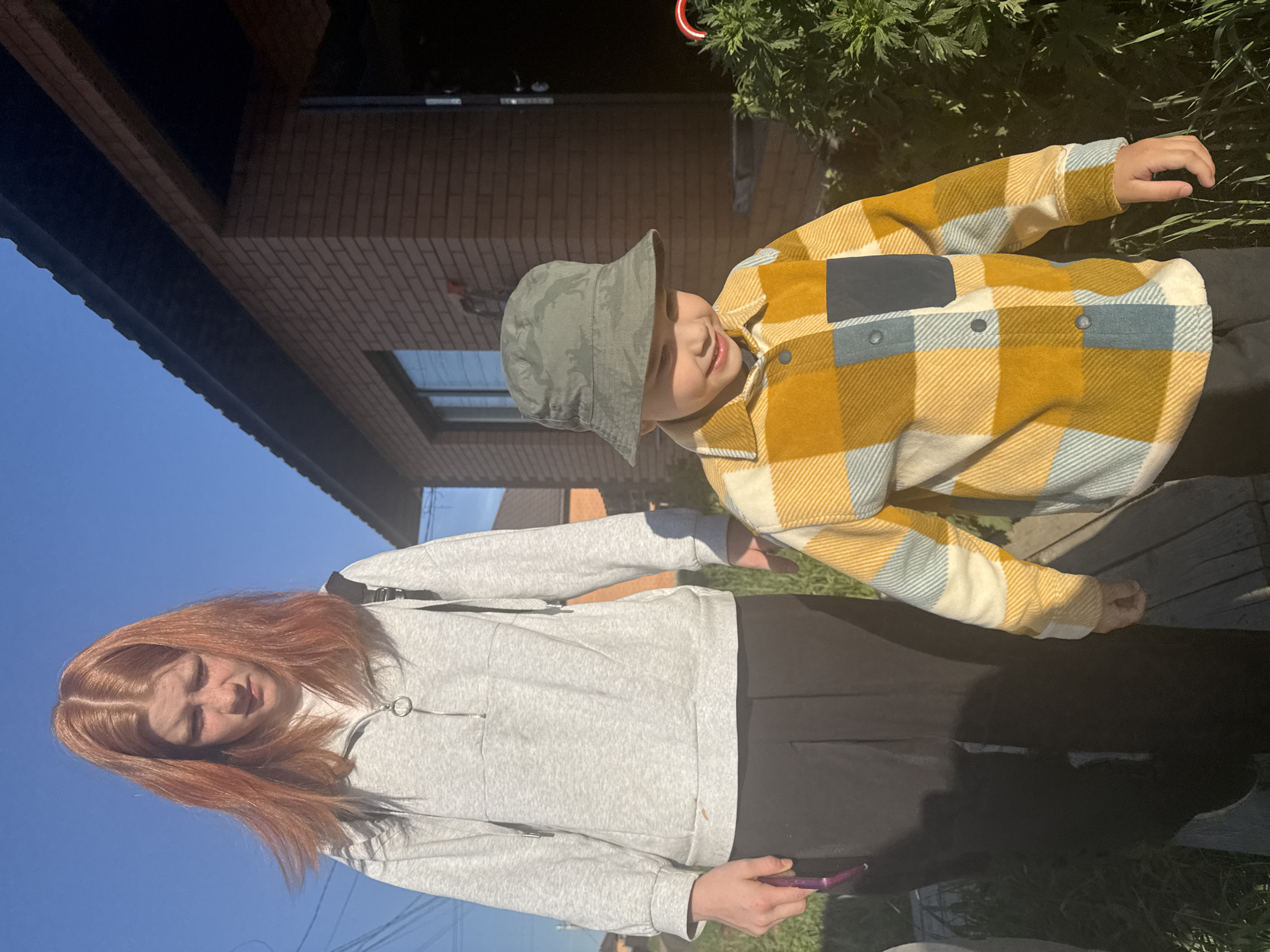 A young woman with red hair, wearing a gray sweatshirt and black pants, stands next to a younger boy in a yellow and light blue plaid shirt and a green bucket hat. The woman is looking down and appears to be holding a cell phone. The boy is looking off to the side with a smile. They are standing outside, near a brick building, possibly a house. Green plants are visible in the background. The sky is blue and sunny. The location is likely Chaikovsky, Russia. The setting appears to be casual and candid.