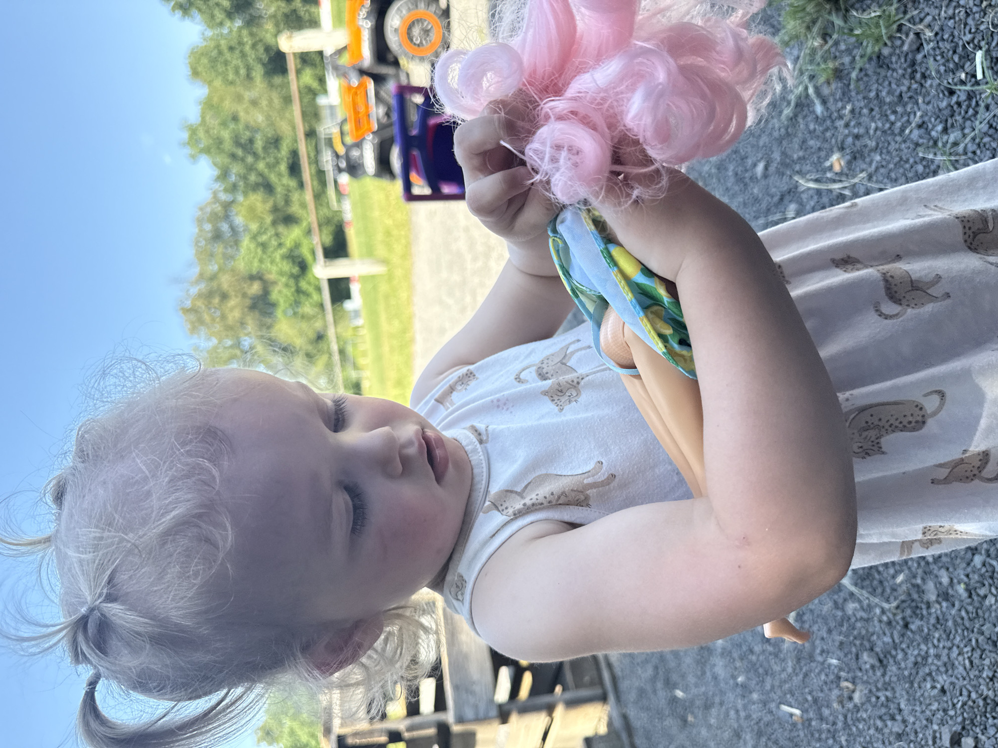 A young girl with blonde hair tied into multiple pigtails plays with a doll outdoors on a sunny day. She is wearing a sleeveless white dress with a pattern of cheetahs. The doll has pink hair and is dressed in a yellow and blue patterned outfit. In the background, there are trees, a fence, and two colorful toy vehicles. The ground is covered with gravel. The setting is implied to be Beltershain, Germany.