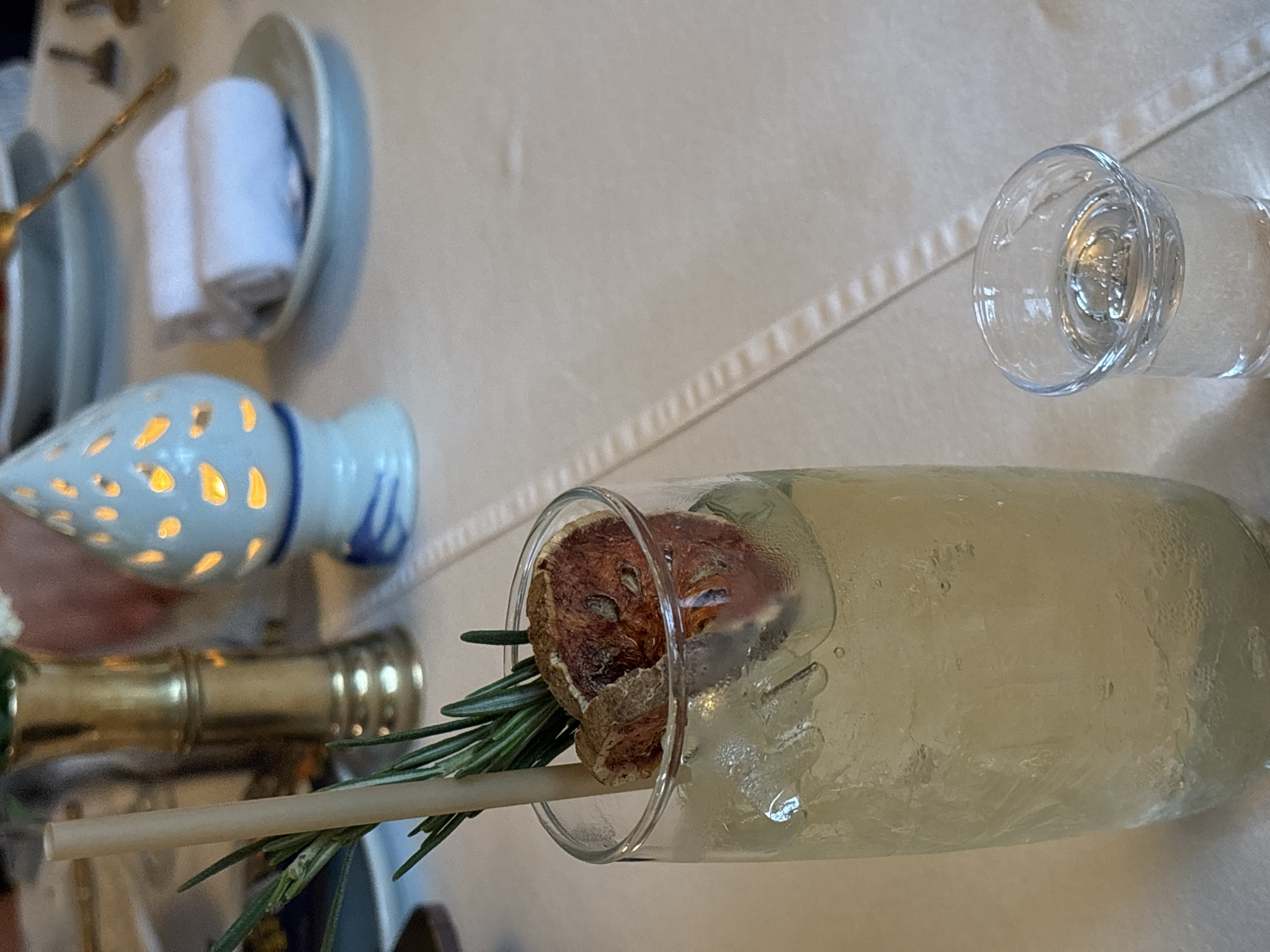 The image shows a table setting in what appears to be a restaurant. The setting includes a glass of pale yellow liquid, presumably a cocktail, with a dried fruit garnish and a sprig of rosemary. A straw is also visible in the glass. Next to the cocktail is a small, clear glass of clear liquid, likely water. Other table settings are visible in the background, including plates, silverware, napkins, and a decorative porcelain object with cutouts.
Without additional information or metadata from the image, it's impossible to confirm the location is Phuket City Municipality, Thailand, or provide a forensic description of the people, actions, or interactions. The objects are detailed as observed in the image.