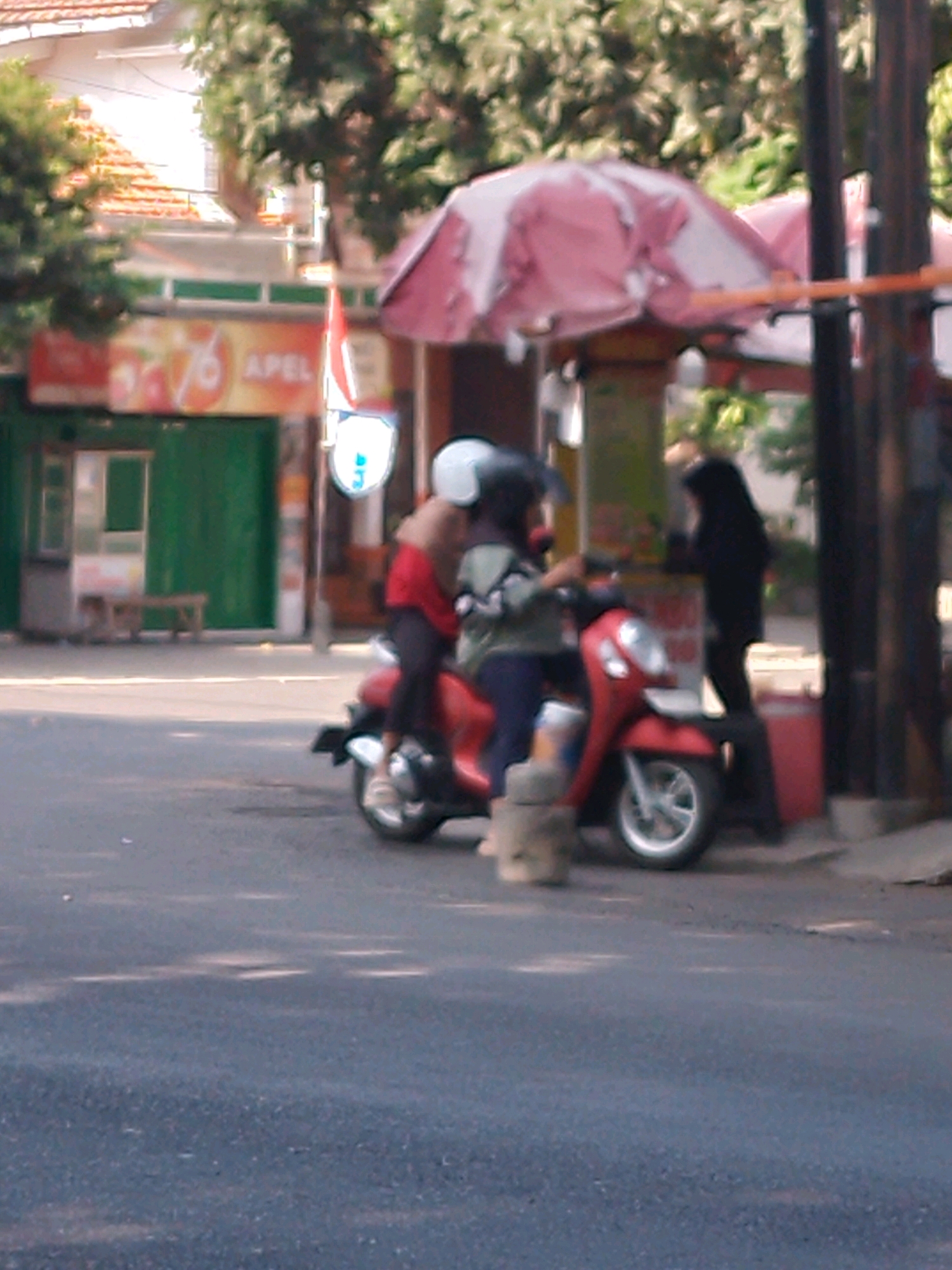 The image depicts a street scene in Kertosono, Indonesia, likely in front of a shop. Two individuals, one dressed in red and the other in a patterned shirt, are seated on a red motorcycle. The person in red, with a white helmet, is in the front, while the person in the patterned shirt, also wearing a helmet, is behind. A third individual, dressed in black and wearing a head covering, stands to the right of the motorcycle. The motorcycle is parked on an asphalt road. In the background, there is a building with a green door and a sign that appears to read "76 APEL". A pink and white awning is visible above the motorcycle. Trees and a wooden bench are also present in the environment. The image is somewhat blurry, suggesting it was taken with a low-resolution camera or in low light conditions.