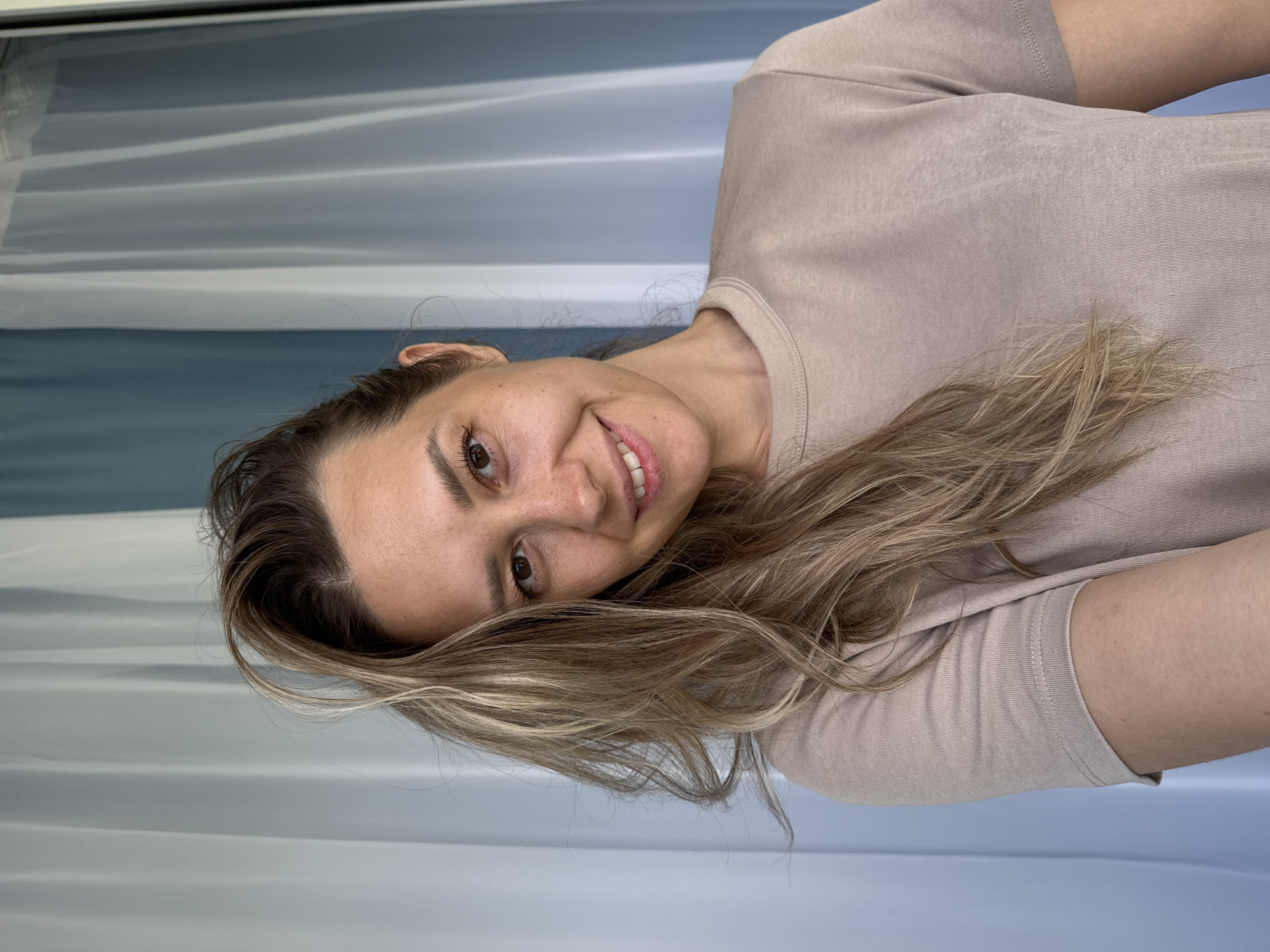 A medium shot shows a woman in her late 20s or early 30s with long, wavy blonde hair, smiling at the camera. She has light skin with freckles and light brown eyes. Her hair is partially covering her face. She is wearing a light brown t-shirt. The background is blurred, showing pale blue and white curtains. The scene appears to be indoors, possibly in a room with a window or balcony, and the lighting suggests daylight. The photo was taken in the city of Nebug, Russia.