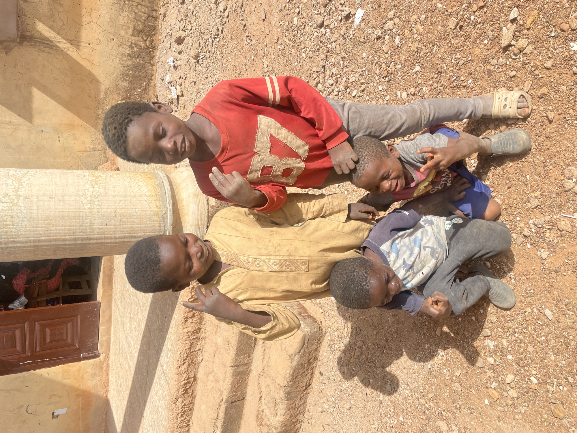 Four young boys are lying on the ground near a building, possibly in Ladura Foron, Nigeria. The ground is reddish-brown and covered with small rocks and gravel. The boy on top is wearing a red shirt with a white number "87" on it and light grey pants with one sandal. Beneath him, another boy in a grey t-shirt and blue shorts is partly visible. The boy beneath him wears a tan garment. The final boy visible is wearing a patterned shirt with grey pants. The boys are close together, possibly interacting playfully. To the left is a portion of a building with visible stairs and a pillar, and a window.
