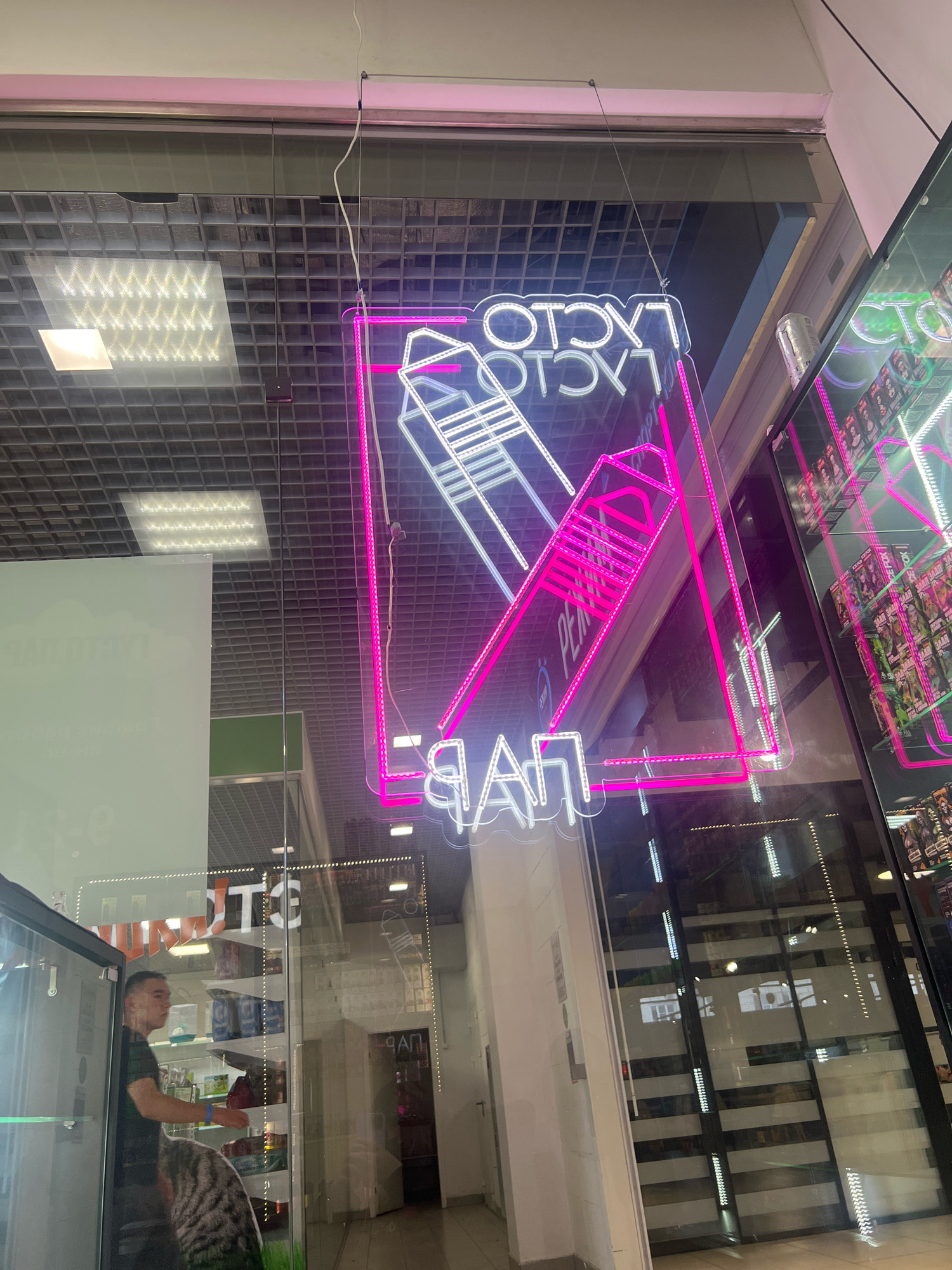 The image shows a storefront with a neon sign that reads "FACTORY" and "PARFUM" in Cyrillic letters. The sign is pink and white and is hanging from the ceiling. The storefront appears to be a retail establishment, possibly a perfume or vape shop, based on the neon sign. Through the glass, shelves with various products are visible, as well as a man in a dark t-shirt looking at the products. The ceiling is made of a grid of square tiles with lights. The exterior of the building is visible as a reflection in the glass, showing a modern facade. The overall impression is that of a brightly lit, contemporary commercial space. Based on the Cyrillic text and context, this image is likely from Nizhny Novgorod, Russia.