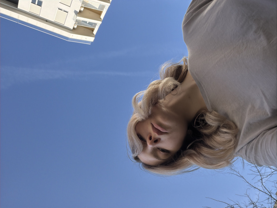 A low-angle upward-facing perspective frames a female individual, positioned in the lower and central foreground. The subject has shoulder-length, wavy, light blonde hair and wears a plain, light-colored (beige or light grey) crew-neck t-shirt. Her head is tilted slightly to her right, and her gaze is directed downward towards the camera. The background consists predominantly of a clear, bright blue sky with a single faint, white contrail visible in the upper center. To the upper right, a section of a multi-story modern building, light-colored with visible windows and balcony structures, is partially visible against the sky. In the lower left quadrant, sparse, bare tree branches are present. The scene is illuminated by natural daylight. This outdoor scene is located in Boreti, Montenegro.
