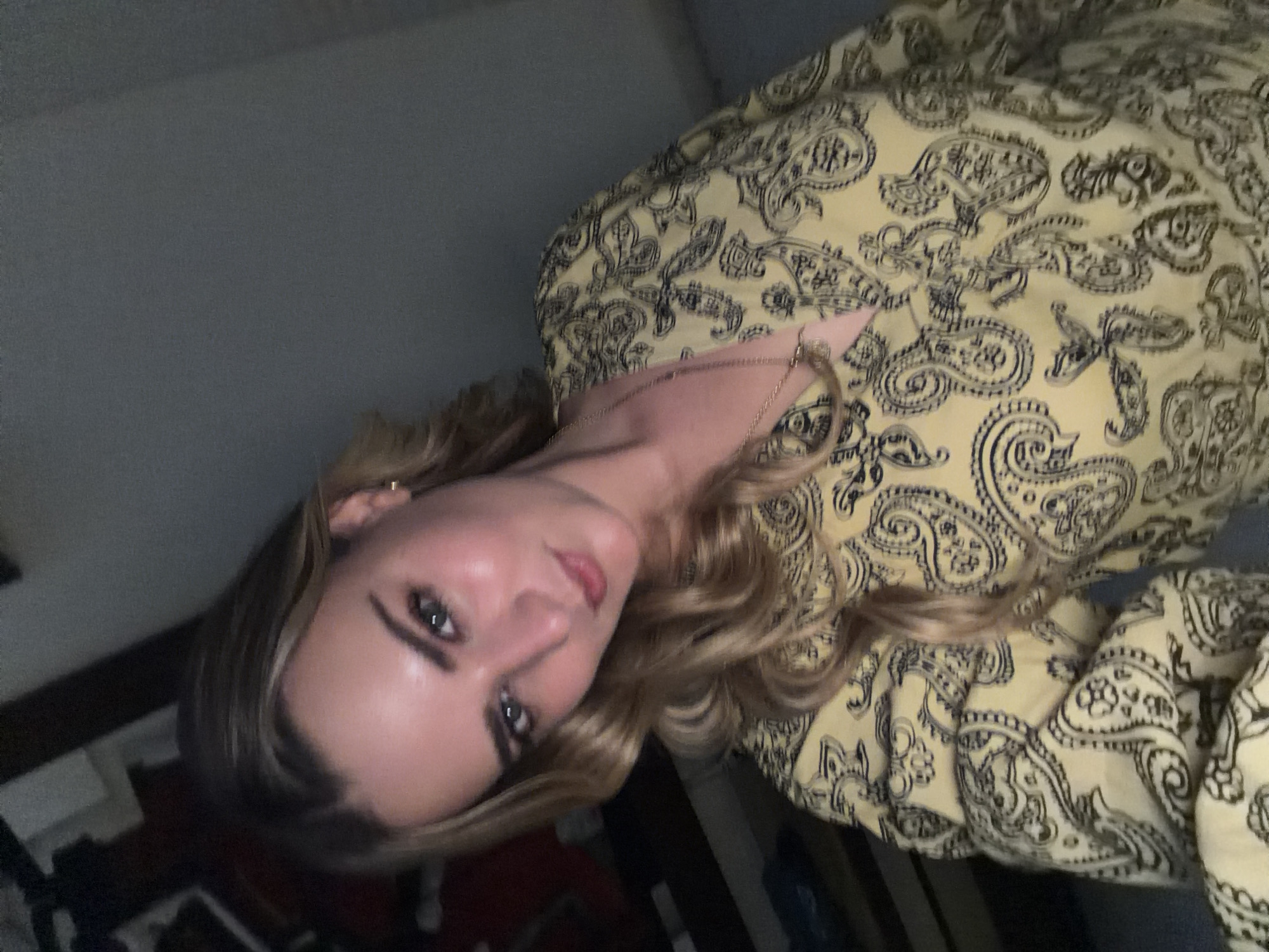 A woman with blond hair and blue eyes is depicted from the shoulders up. She is wearing a yellow top with a black paisley pattern. The background is a dark gray, possibly a wall or other interior feature. The location is unknown.