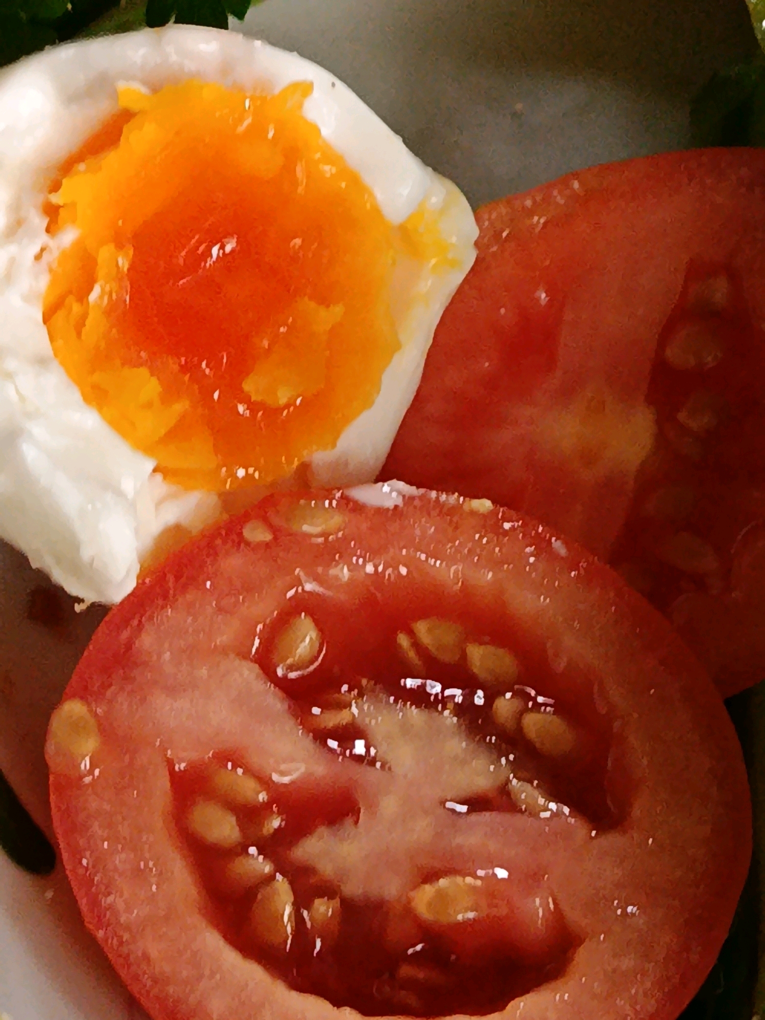 A close-up, overhead shot shows a sliced tomato and a soft-boiled egg. The tomato slices are bright red and juicy, with visible seeds and water droplets on their surface. The soft-boiled egg has a bright orange yolk that is still liquid and a white, somewhat wispy albumen. The background is blurred but appears to be a dark surface, possibly a plate or bowl. There are no people or any indication of a specific location.