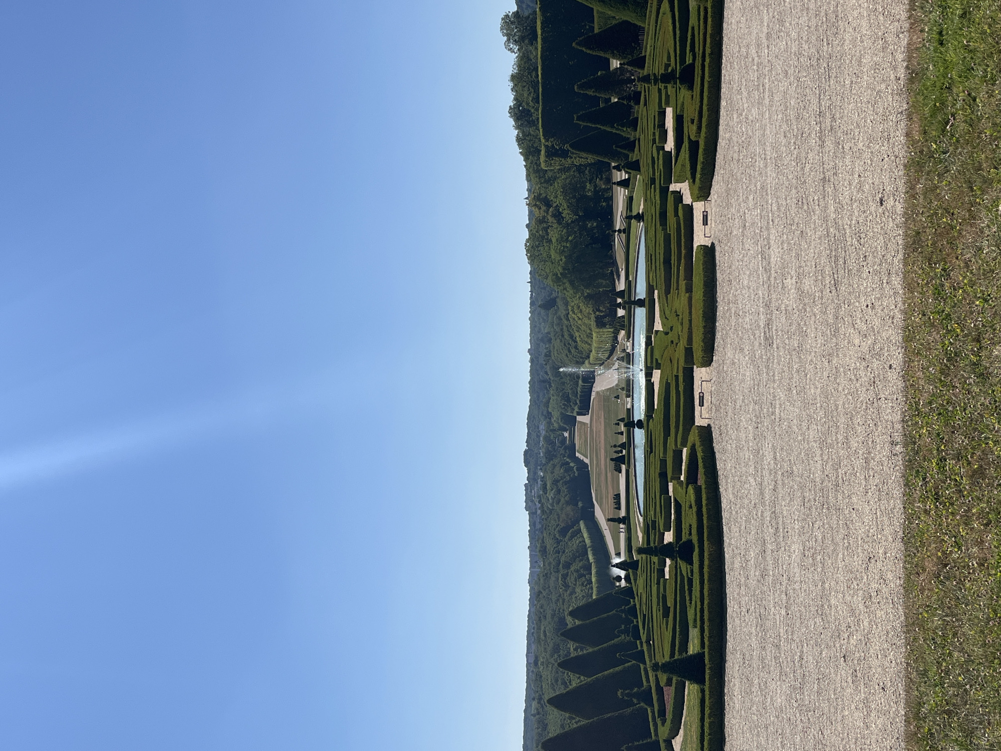 This is a photograph of the Parc de Sceaux in Sceaux, France, taken on a clear, sunny day. The image is dominated by the expansive formal gardens, featuring meticulously sculpted hedges and manicured trees arranged in geometric patterns. In the foreground, a gravel pathway stretches across the bottom half of the frame, bordered by a strip of green grass. Beyond the gravel, a large, circular fountain is visible, its water jets reaching upwards. The garden then extends into a vast, tree-lined parkland, with a long, straight canal or body of water stretching towards the horizon. The canal is flanked by rows of trees, and a distant line of buildings can be seen on the horizon, hinting at the city beyond the park. The sky above is a brilliant, cloudless blue. There are no people visible in the frame.