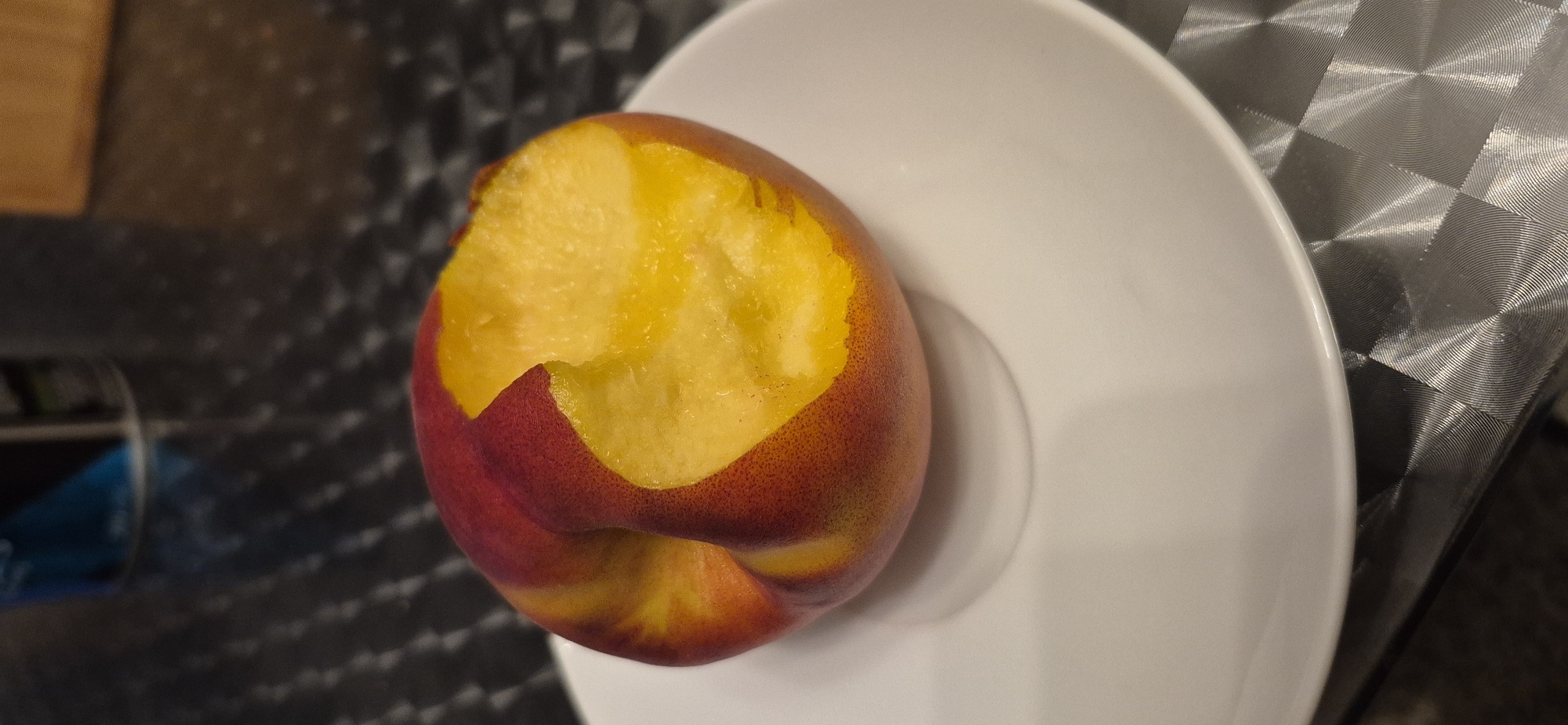 A nectarine with a bite taken out of it rests on a white plate. The nectarine is primarily red and yellow, with the flesh being a bright yellow. The plate is white and sits on a dark, patterned surface. In the background, a blurry can is visible, and a wooden surface is also out of focus. The scene is indoors, and no specific location in Baden-Baden, Germany, can be identified from the image.