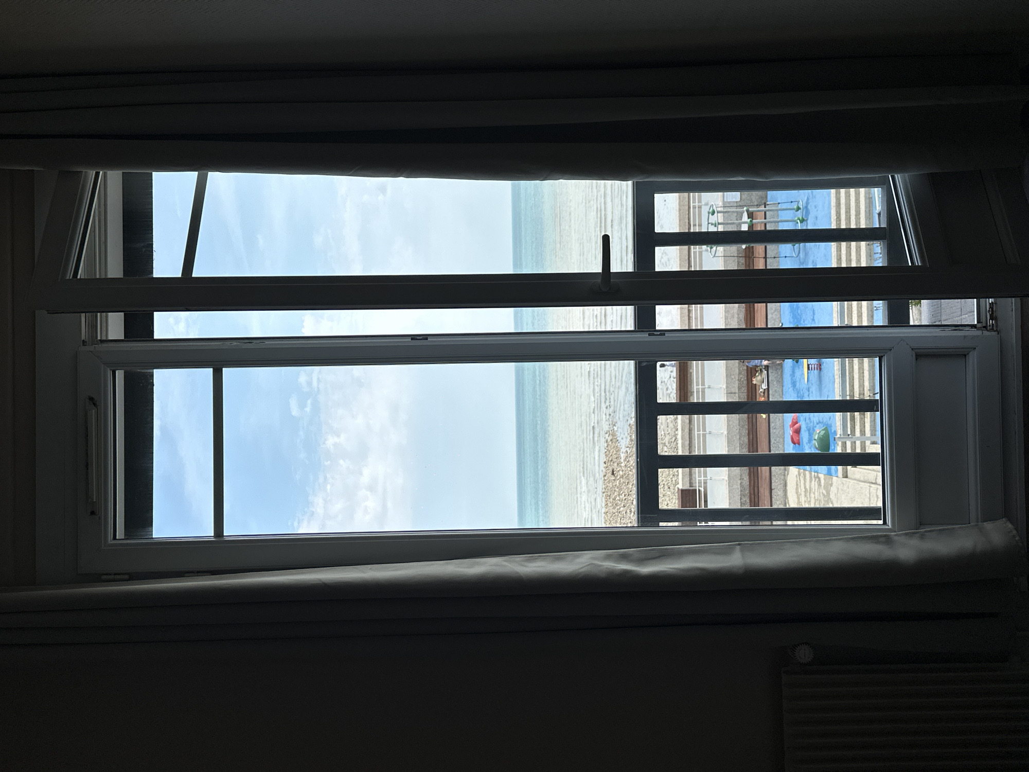 This image captures a view through an open French door, looking out towards the sea from a room in Saint-Valery-en-Caux, France. The foreground is dominated by dark gray curtains on either side, framing the doorway. To the left, partially visible, is a radiator. The open door reveals a second layer of glass panels with a black railing in front of them. Beyond the railing, a vibrant blue surface, likely a playground, is visible with red and green play structures. Further out, the ocean stretches to the horizon under a pale blue sky with scattered clouds. The shoreline is rocky, with a narrow strip of pebbles meeting the water. The overall impression is one of a tranquil seaside scene viewed from an interior space.