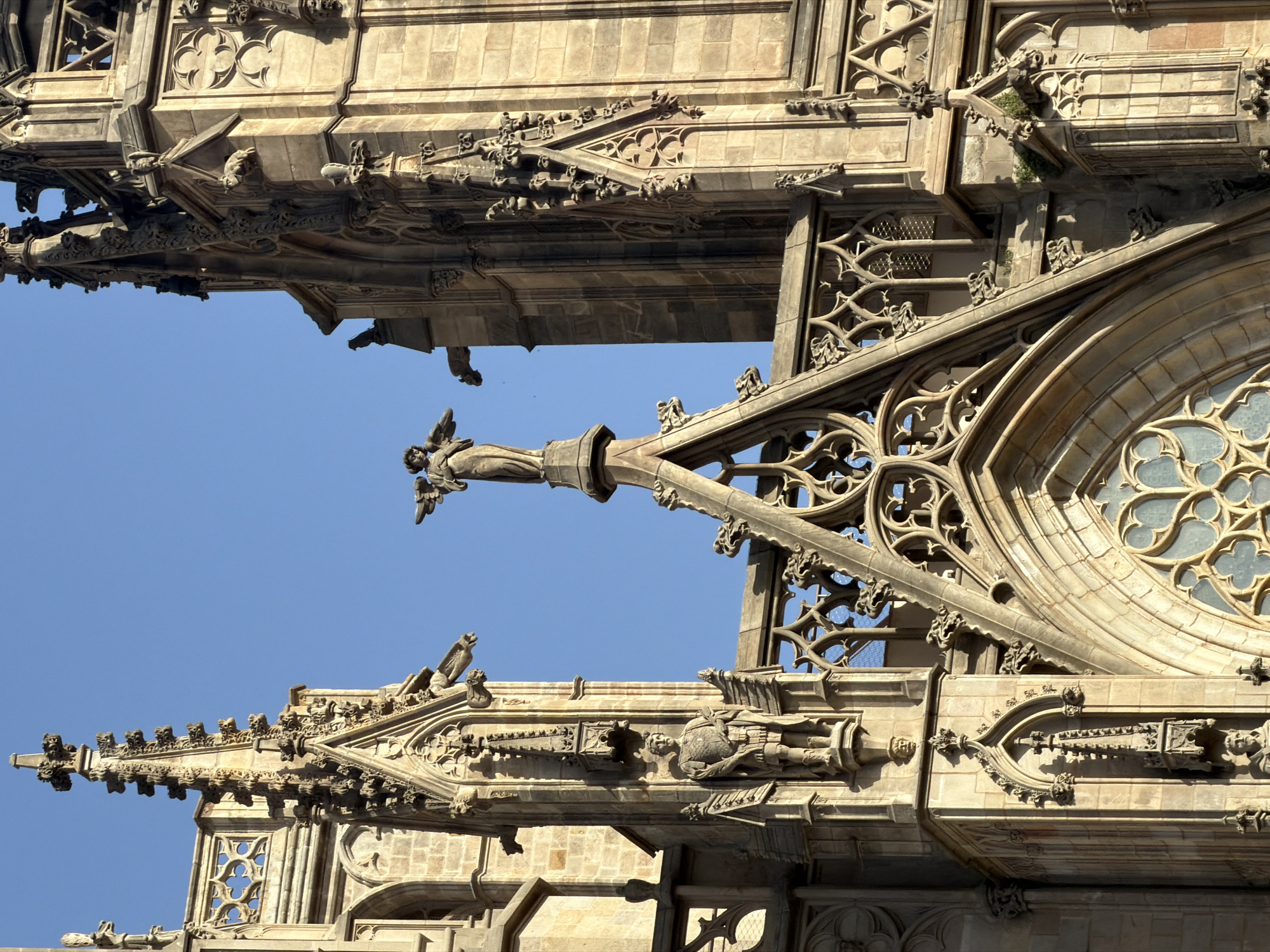 The image displays the upper sections of the Barcelona Cathedral, a Gothic cathedral located in the heart of the Gothic Quarter of Barcelona, Spain. The architectural style is characterized by intricate stonework, pointed arches, and spires. Prominently featured is a statue of an angel at the apex of a spire, with outstretched wings. To the left, another statue depicts a figure in armor. Gargoyles are visible as architectural embellishments on the facade. The sky above is a clear, bright blue. The stonework exhibits the warm, sandy tones typical of aged sandstone or limestone. The overall scene captures the grandeur and detailed craftsmanship of Gothic architecture against a serene sky.