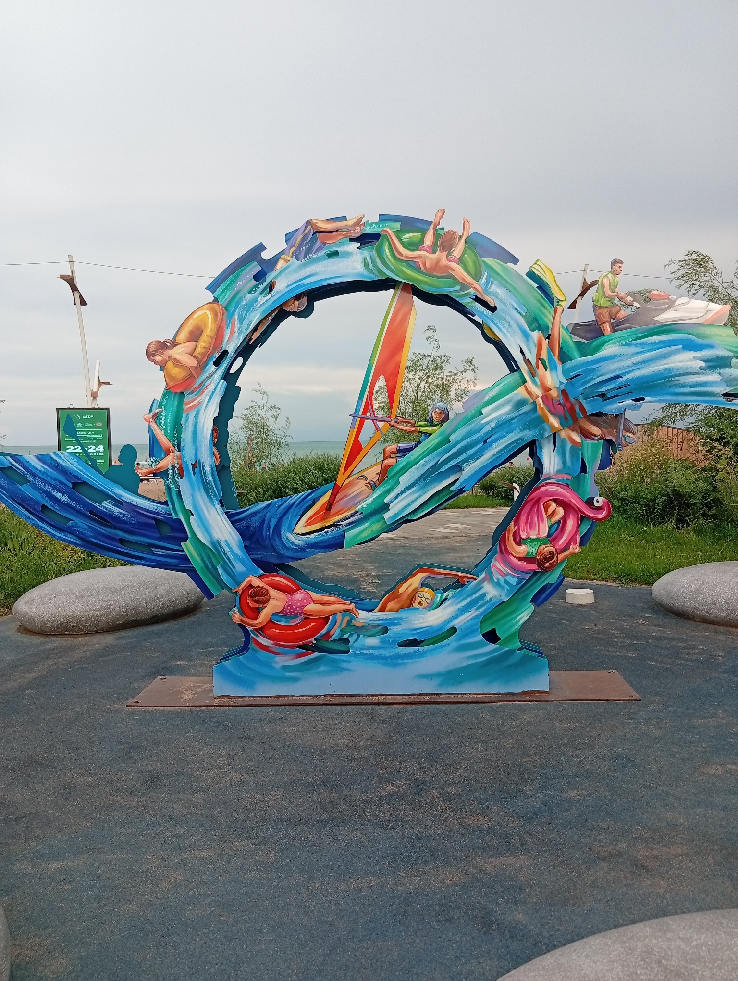 This is a colorful, outdoor sculpture depicting people enjoying various water activities. The sculpture is circular in shape, resembling a wave or whirlpool, with stylized figures of swimmers, a person on a jet ski, and a windsurfer integrated into the design. The sculpture is set on a dark blue rubberized surface, with two large gray stones flanking its base. In the background, there is a body of water, greenery, and a cloudy sky. A digital display board with green text and numbers is visible to the left of the sculpture. The scene appears to be in a public park or recreational area.