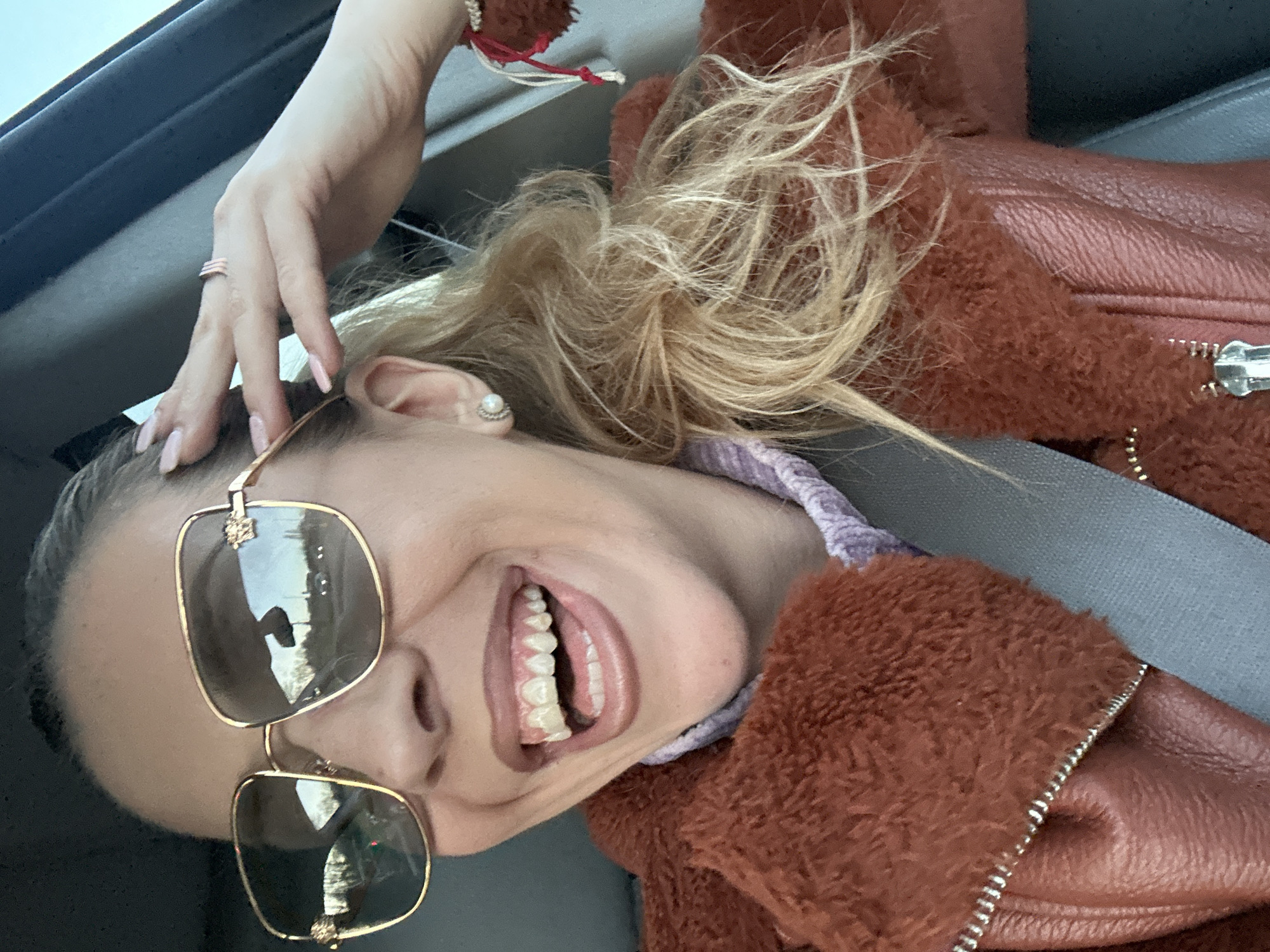A blonde woman in a car wearing a brown fur-lined jacket, a seatbelt, and large square sunglasses is seen smiling at the camera. She is wearing a ring on her left hand, which is raised to the top of her head. It is not possible to determine her location in the city of Basildon, United Kingdom, based on the image.