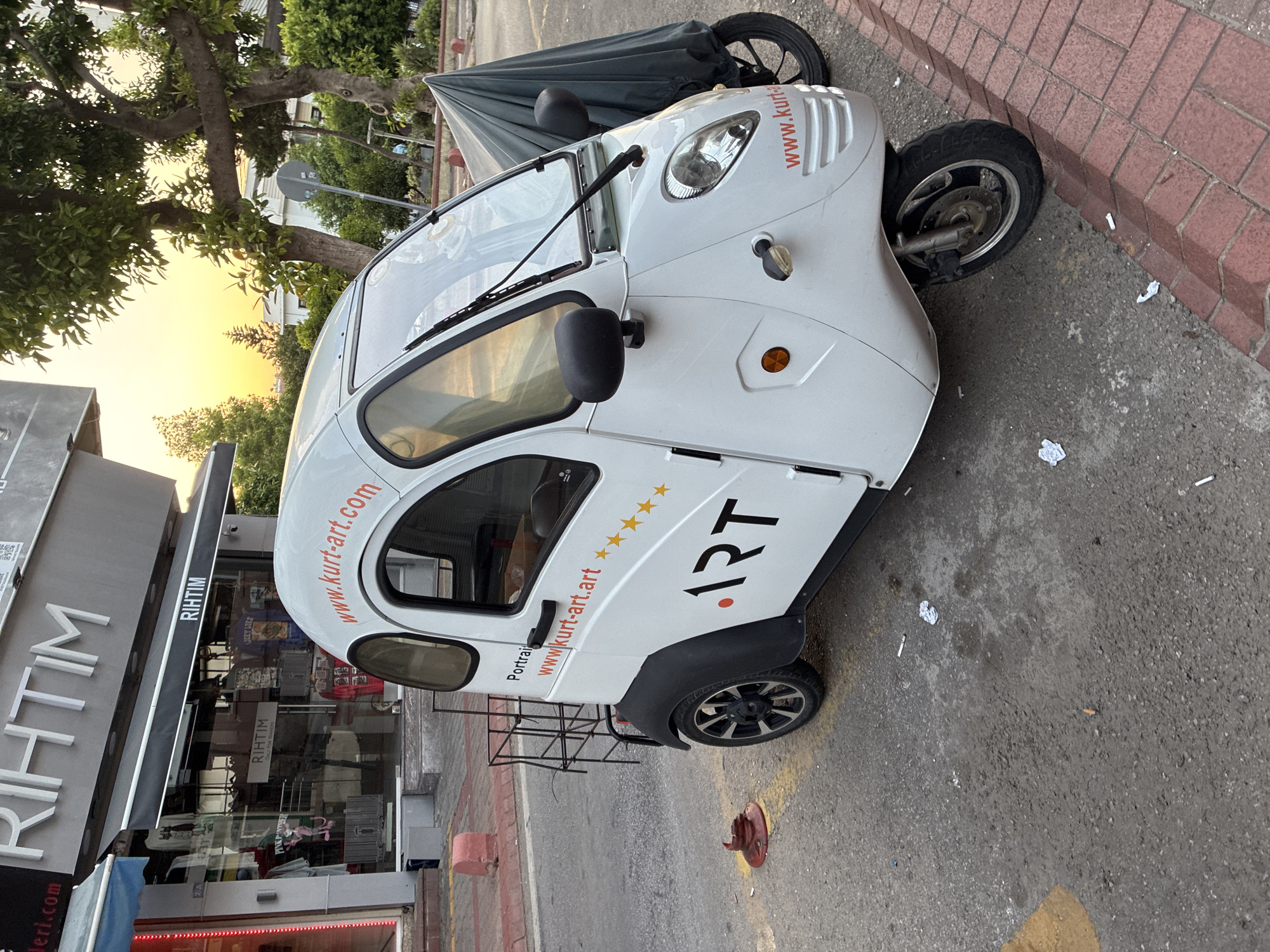 A white, three-wheeled electric scooter is parked on a sidewalk in front of a storefront. The scooter has a covered rear section and a small, enclosed passenger cabin with a windshield and wipers. The storefront has a large sign that reads "RIHTIM" in large silver letters, and the windows display clothing items. The scooter has "www.kurt-art.com" printed in orange on both sides and the front. The surrounding environment includes trees, a brick-lined sidewalk, and a paved road. The sky appears to be lit with the soft light of sunset. The scene is likely in Alanya, Turkey, given the store's signage.