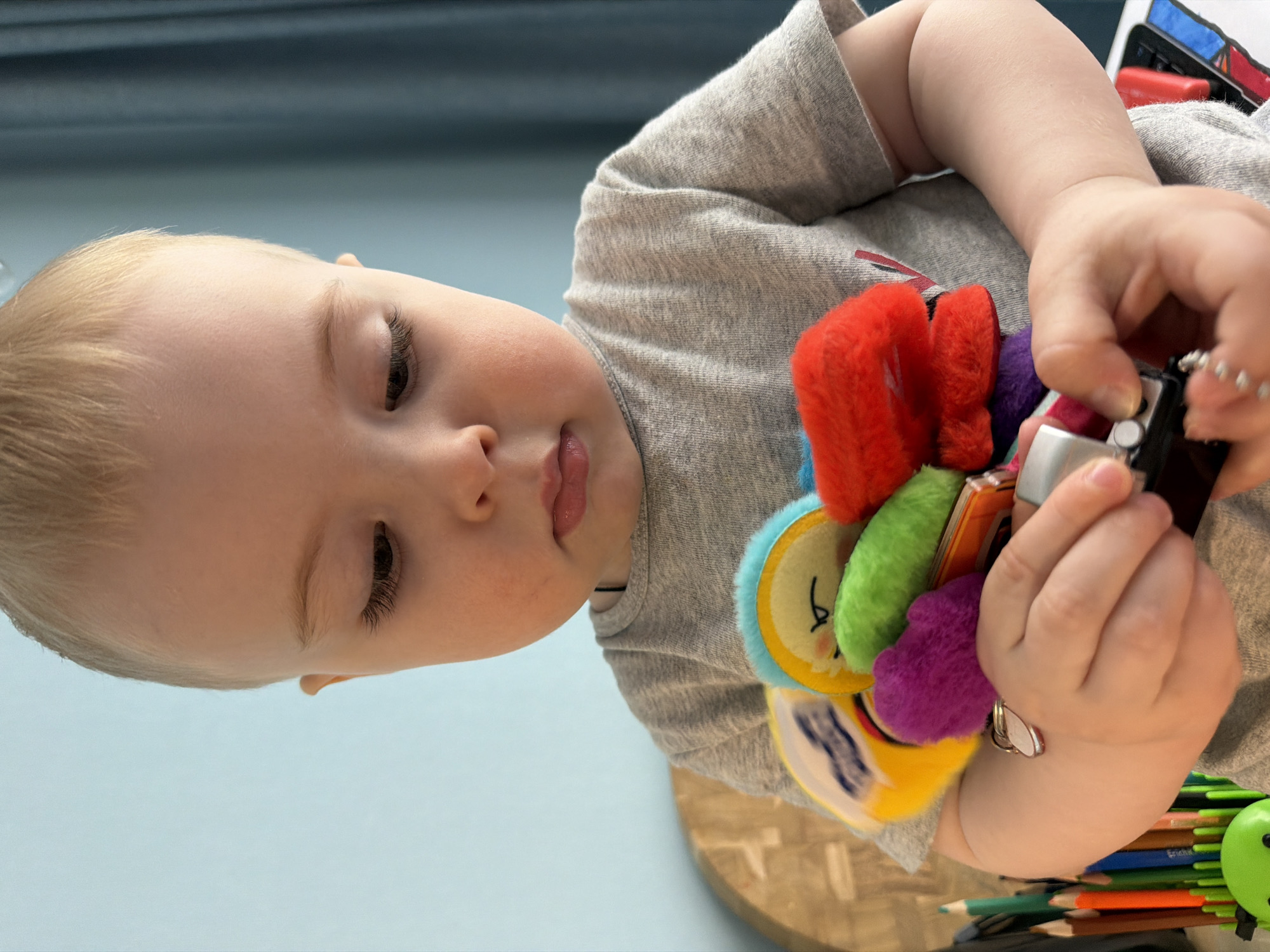A young child with fair skin and blond hair, wearing a gray t-shirt, is shown holding a colorful toy consisting of multiple soft, textured shapes and a small, black object that appears to be a toy car or electronic device. The child is looking down at the toy. Several colored pencils or crayons are visible in the lower right corner of the image. The background is a light blue surface. The image does not show any information that would indicate a location in Votkinsk, Russia. The setting appears to be indoors, possibly a home or daycare environment.