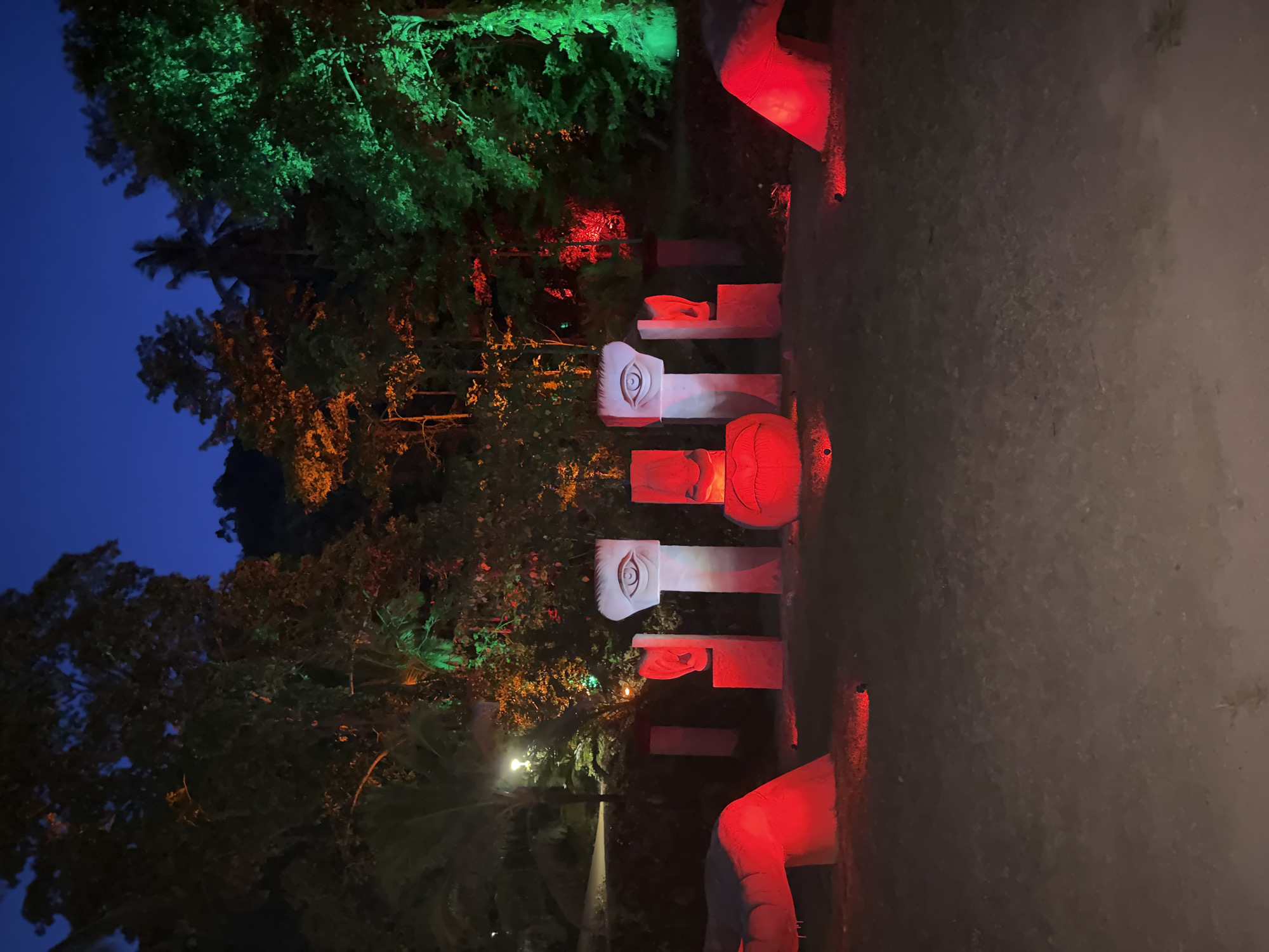 This outdoor scene in Baan Lipa Noi, Thailand, depicts an art installation at night. A series of painted, concrete statues with eyes and facial features are illuminated with red lights. Nearby trees are lit with green and orange lights, creating a colorful contrast. The background shows a dark blue sky, and the ground appears to be a dirt path.